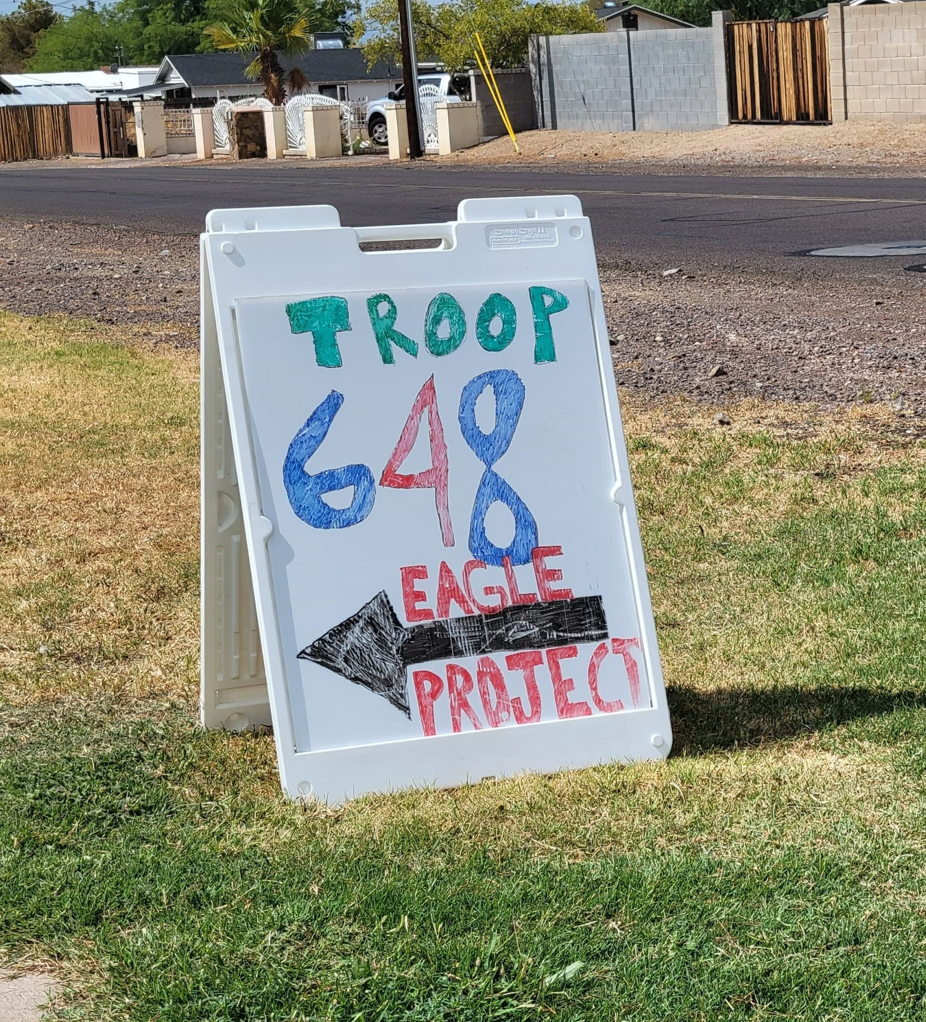 Hand-lettered white A-frame sign reading TROOP 648 EAGLE PROJECT with a black arrow, standing on grass beside a desert neighborhood street
