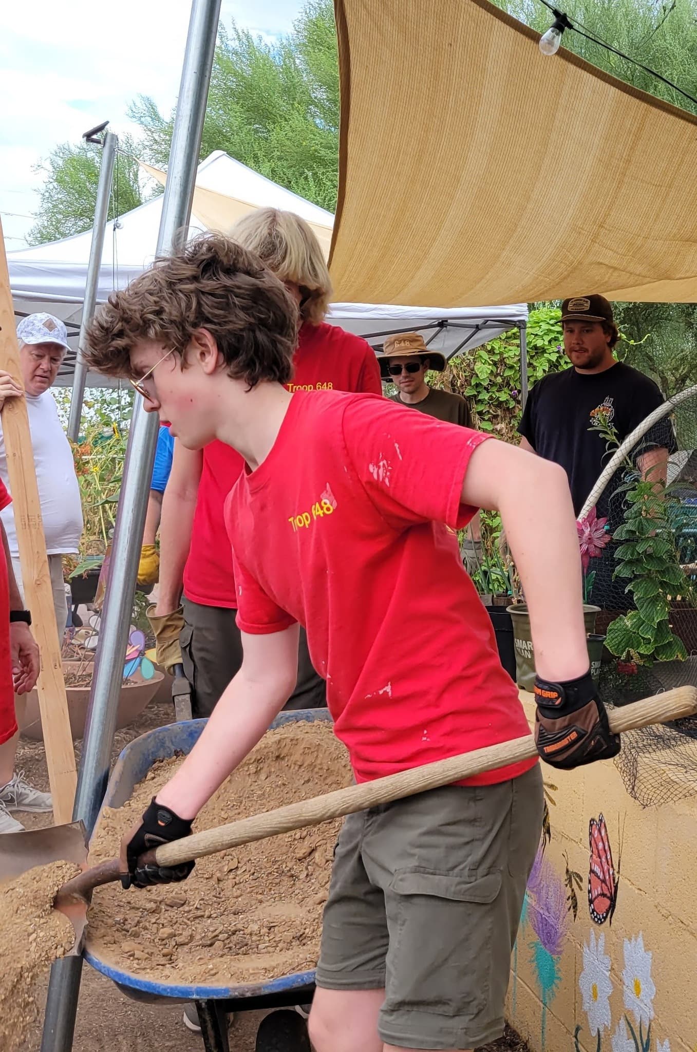 Teen in a red troop shirt shovels gravel from a blue wheelbarrow under a tan shade sail; a butterfly and flowers are painted on the block wall