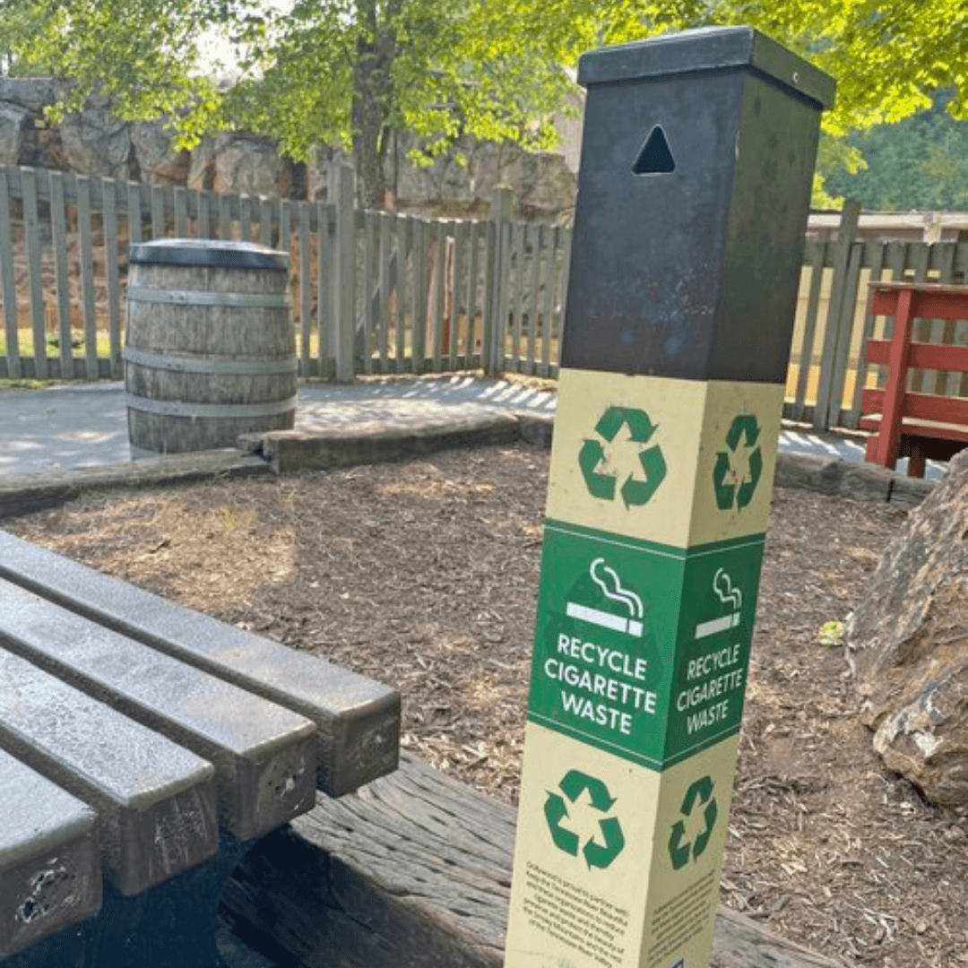 Cream and green receptacle labeled RECYCLE CIGARETTE WASTE with recycling arrows, next to a wooden bench and barrel on a mulched path