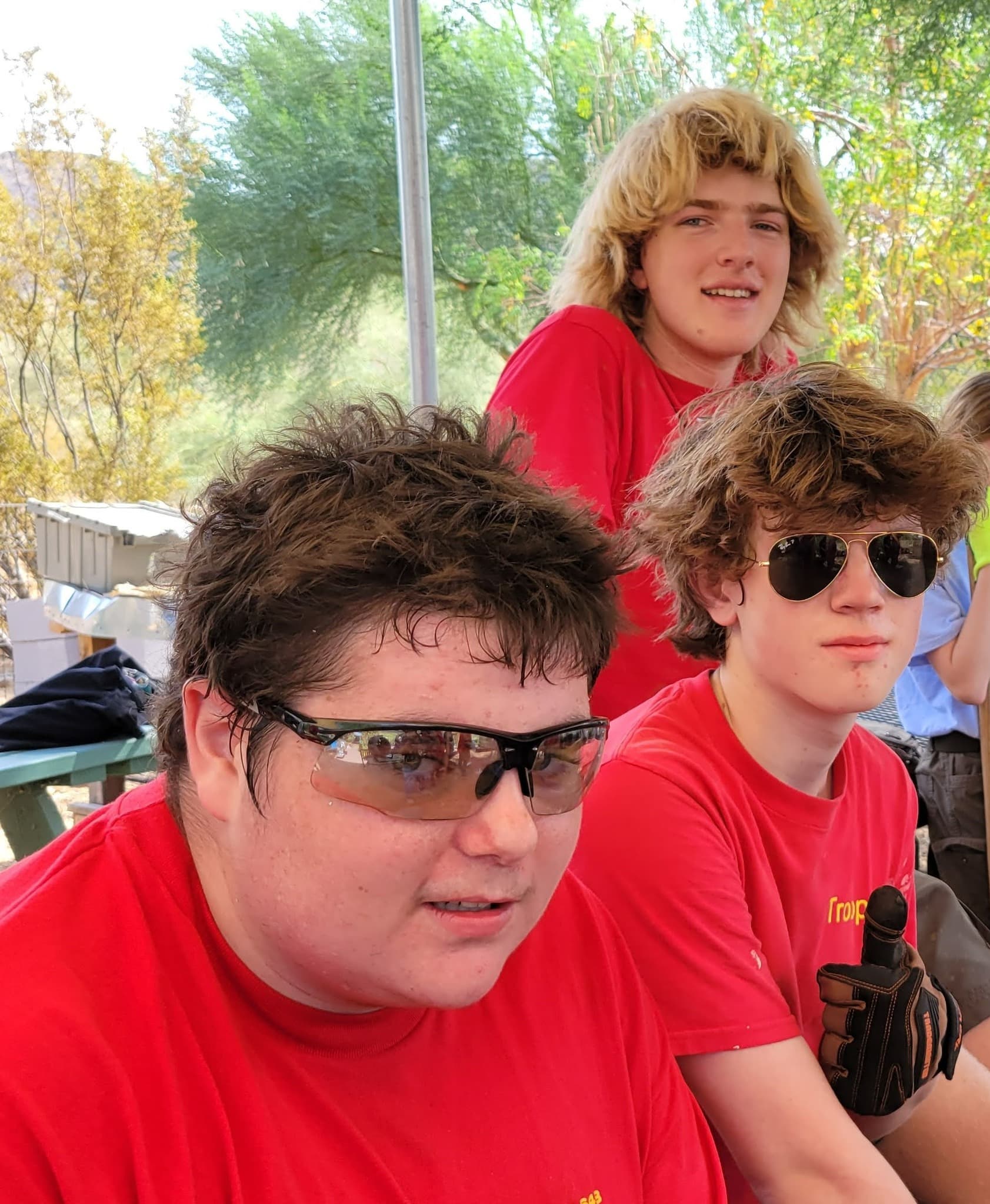 Close-up of three teenage boys in red troop shirts and sunglasses resting at a picnic table under desert trees