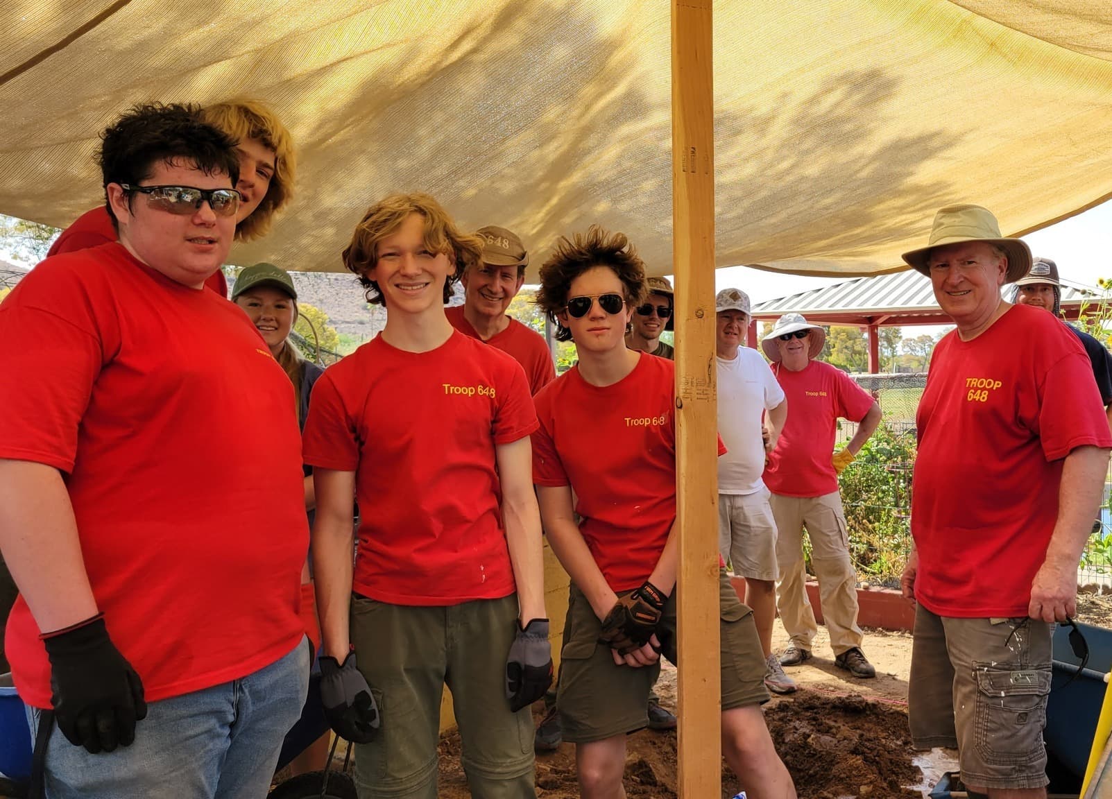 Volunteers in red troop T-shirts gathered under a tan shade canopy beside a freshly dug garden bed, an older man in a wide-brim hat at right