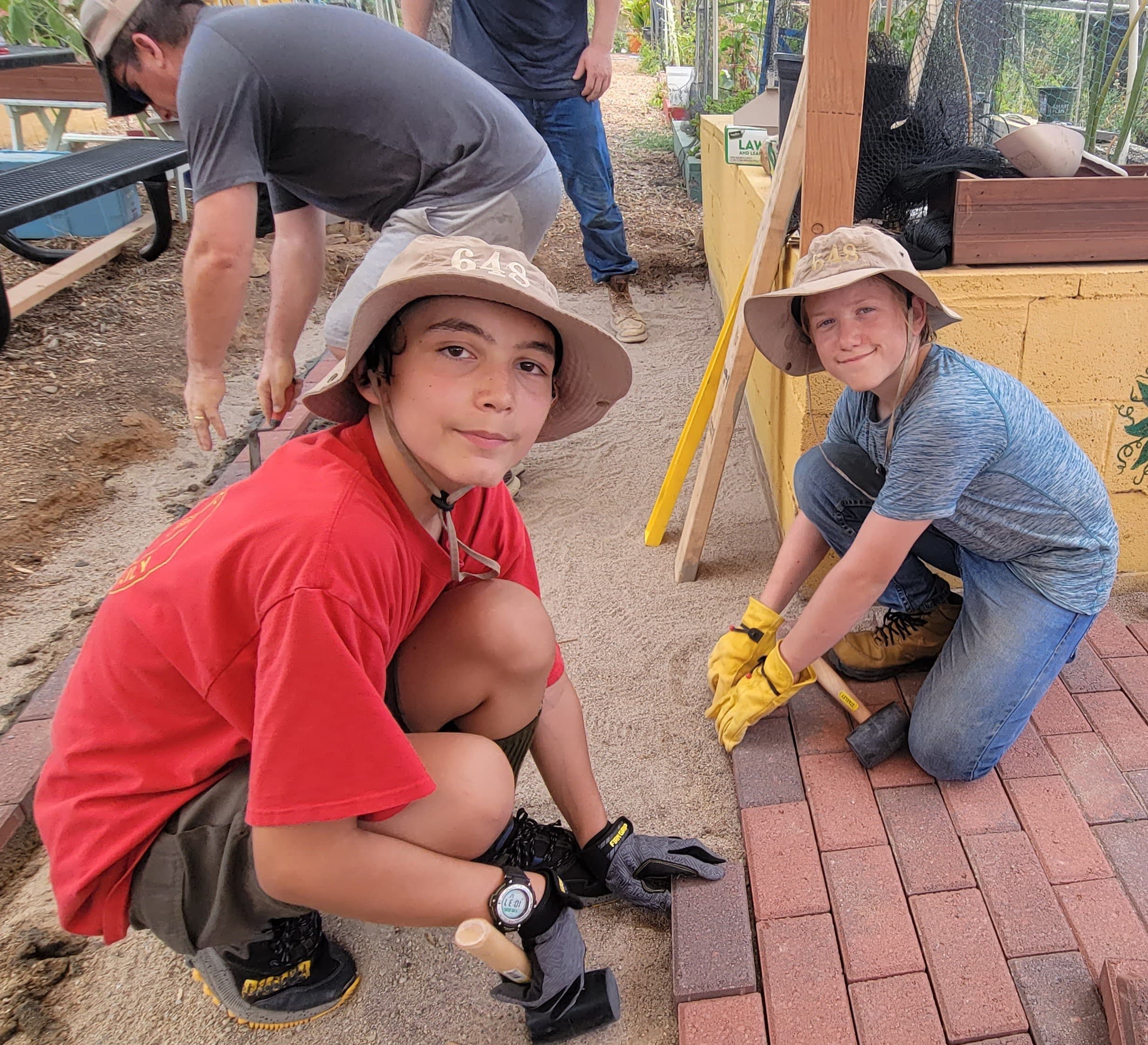 Two boys in bucket hats crouch on sand laying red pavers, one holding a rubber mallet in yellow gloves, an adult troweling behind them