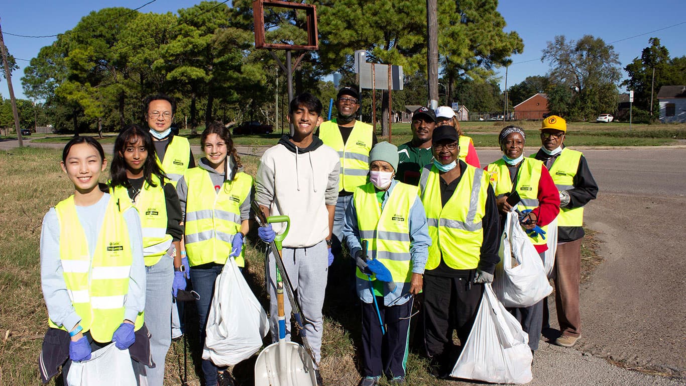 A dozen volunteers of mixed ages in yellow high-visibility vests crowd together for a group photo on a grassy verge, holding white collection bags, shovels and litter grabbers.