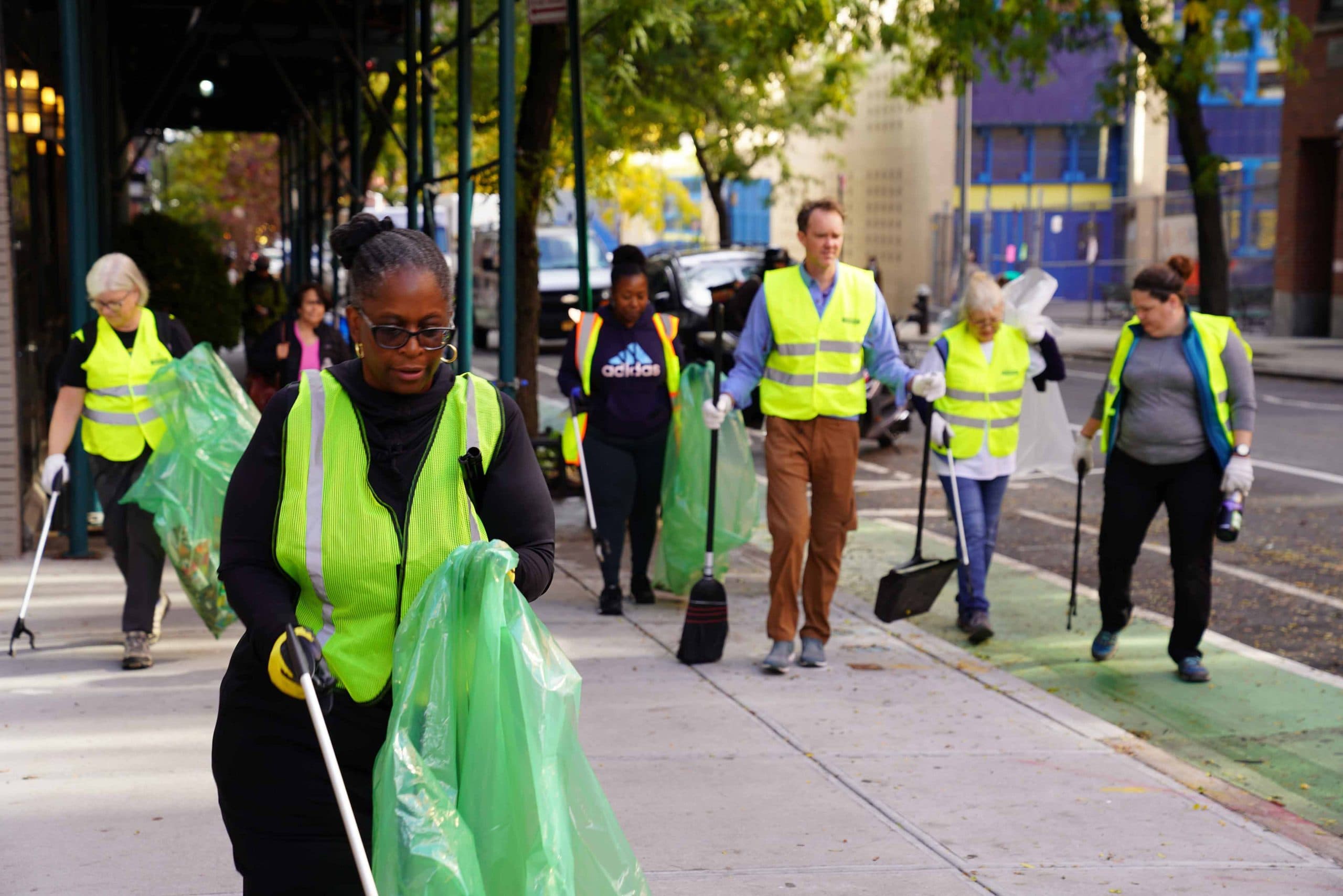 Six volunteers in high-visibility vests work their way along a city sidewalk under an elevated rail bridge, carrying green collection bags and litter grabbers, a woman in the foreground holding hers open mid-sweep.