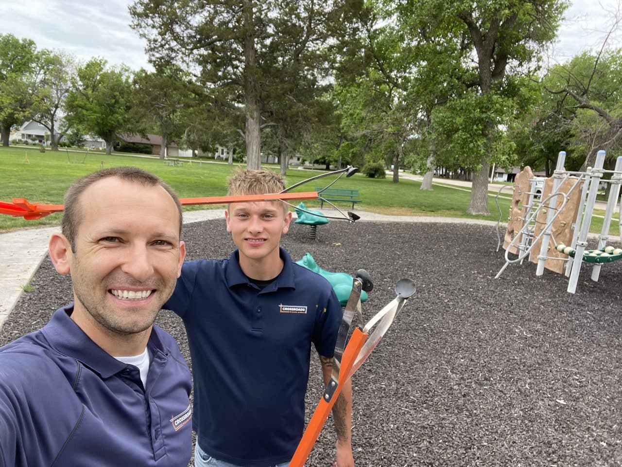 Two Keep Lexington Beautiful volunteers in Crossroads polos take a smiling selfie holding orange litter grabbers at a park playground.