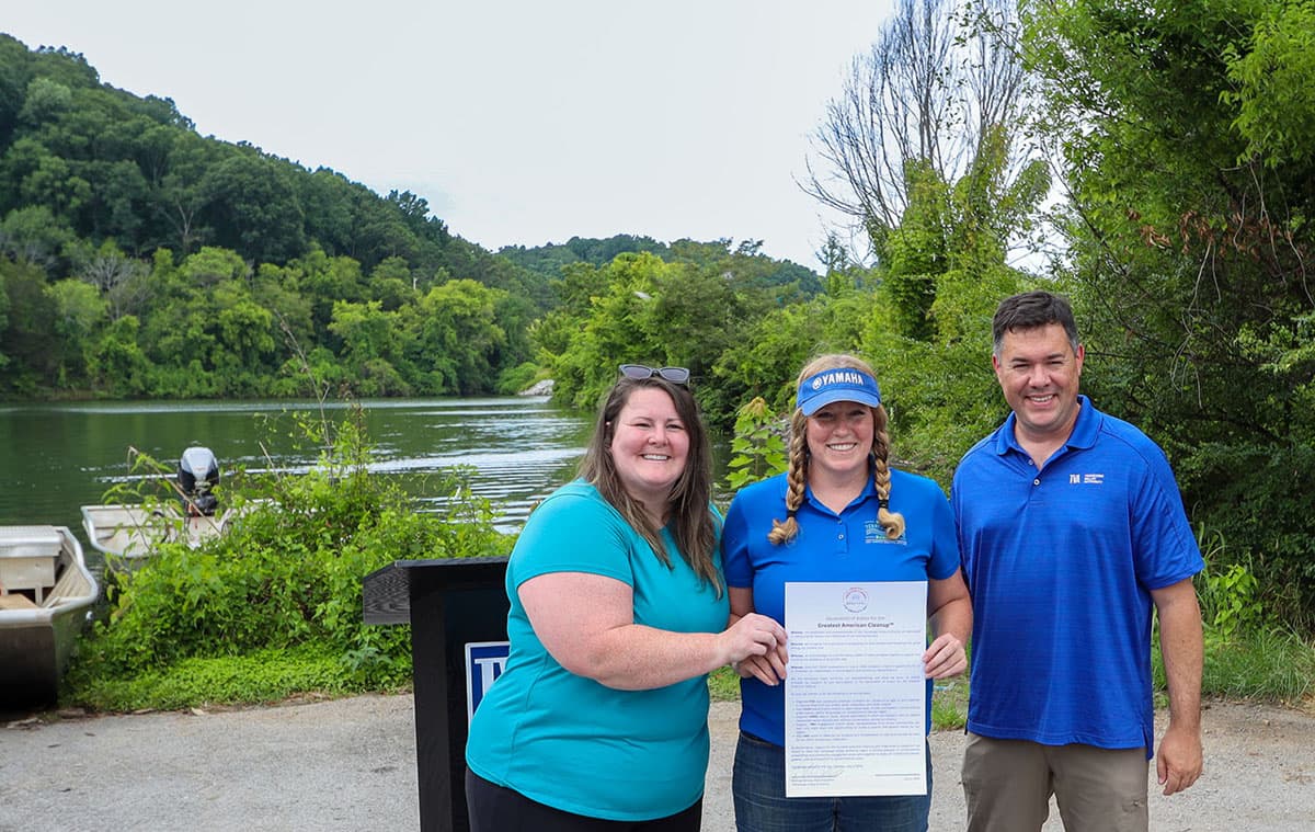 Three people stand on a gravel boat ramp beside a wide green river, holding a printed Greatest American Cleanup proclamation between them. A woman in a teal t-shirt with sunglasses pushed into her hair is on the left, a woman in a blue polo and blue Yamaha visor with long plaits holds the document in the centre, and a man in a blue polo and khaki trousers stands on the right. A small aluminium boat and a wooded hillside are behind them.