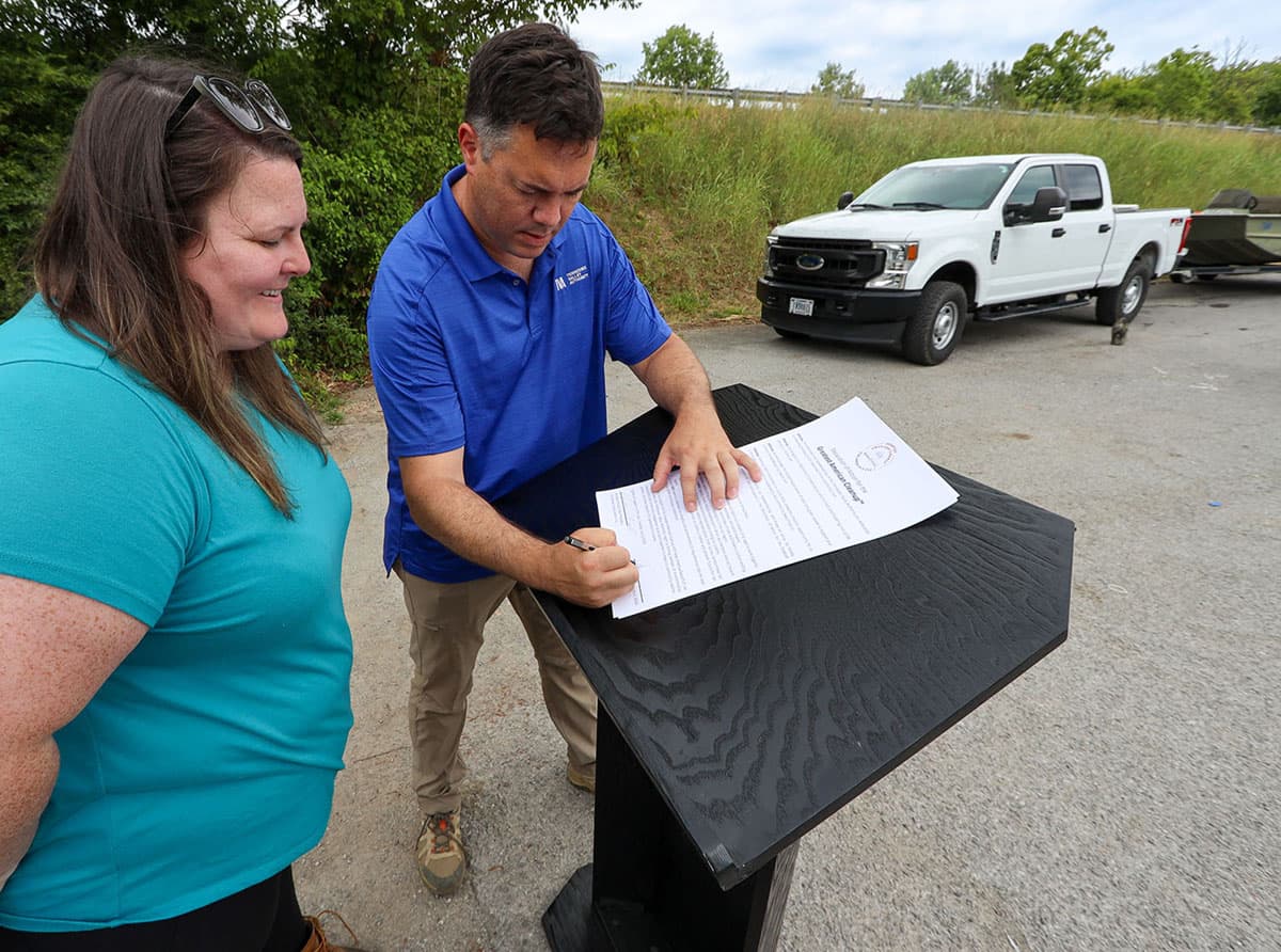 A man in a blue polo shirt leans over a black wooden lectern on a car park apron, pen in hand, signing a printed Greatest American Cleanup proclamation. A woman in a teal t-shirt with sunglasses in her hair watches from the left. A white pickup truck with a trailer is parked on the grass verge behind him.