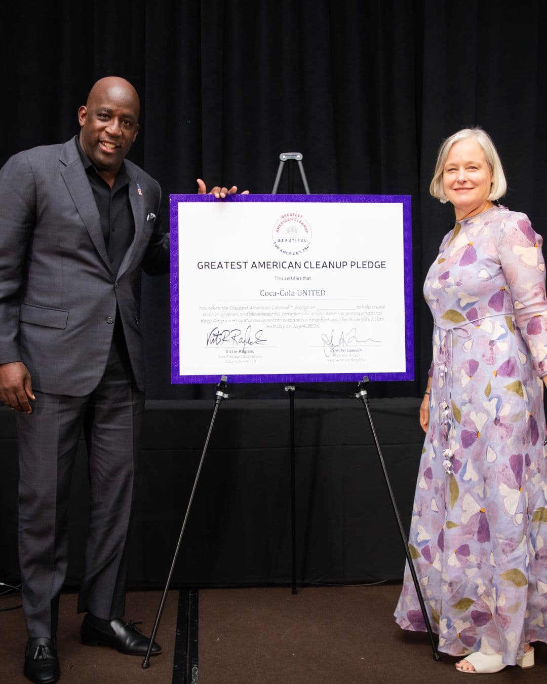 Two people stand either side of a large framed certificate on a black easel, reading GREATEST AMERICAN CLEANUP PLEDGE and certifying Coca-Cola UNITED, signed by Victor Ragland and Jennifer Lawson. A man in a charcoal suit rests a hand on the top corner of the board on the left; a woman in a pale lilac floral dress stands on the right. The backdrop is black stage drapery.