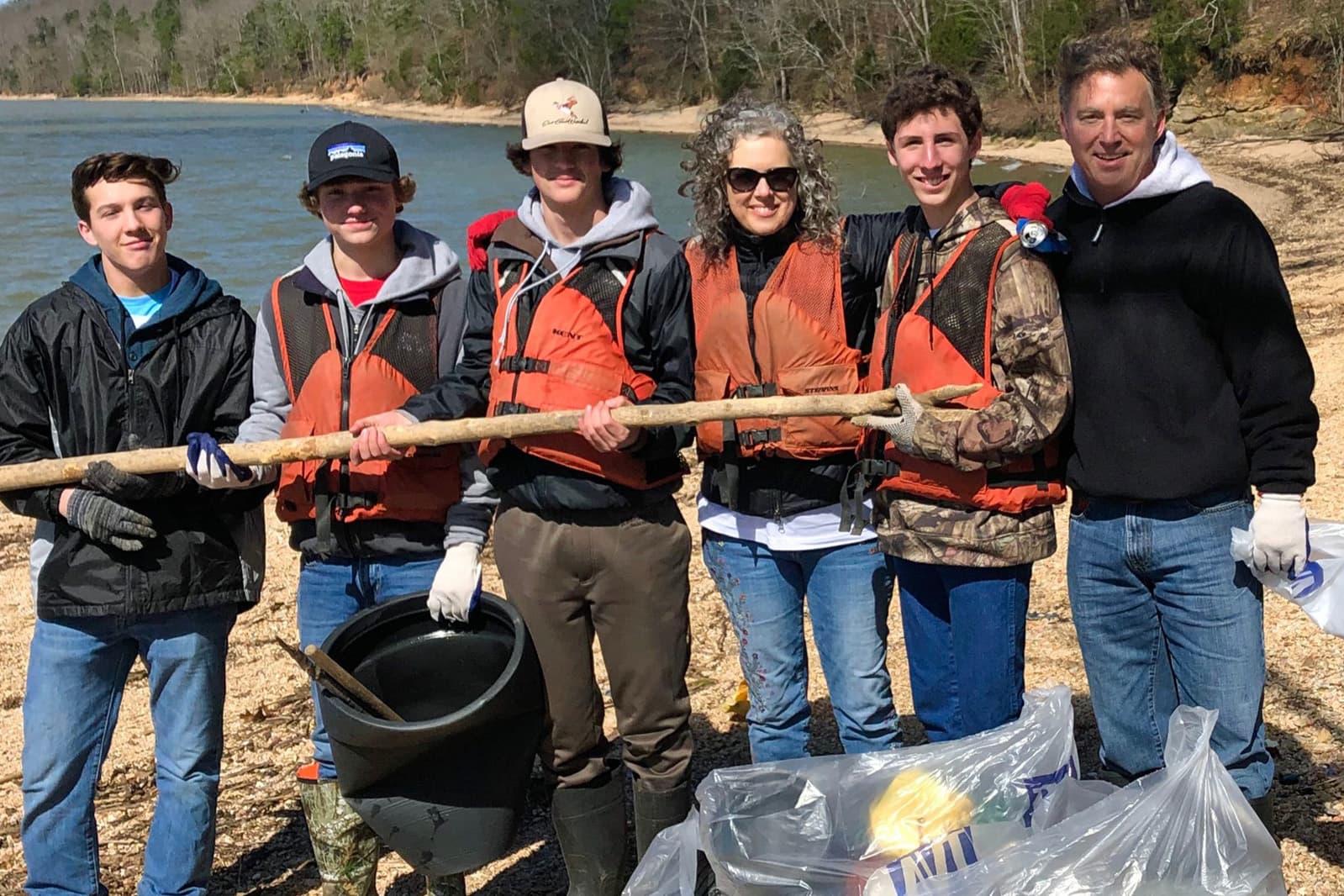 Six people stand on a pebbled lake shore in front of dark green water and bare winter trees, four wearing orange buoyancy vests, holding a long weathered wooden pole between them. A black bucket and a bulging clear rubbish bag sit at their feet.