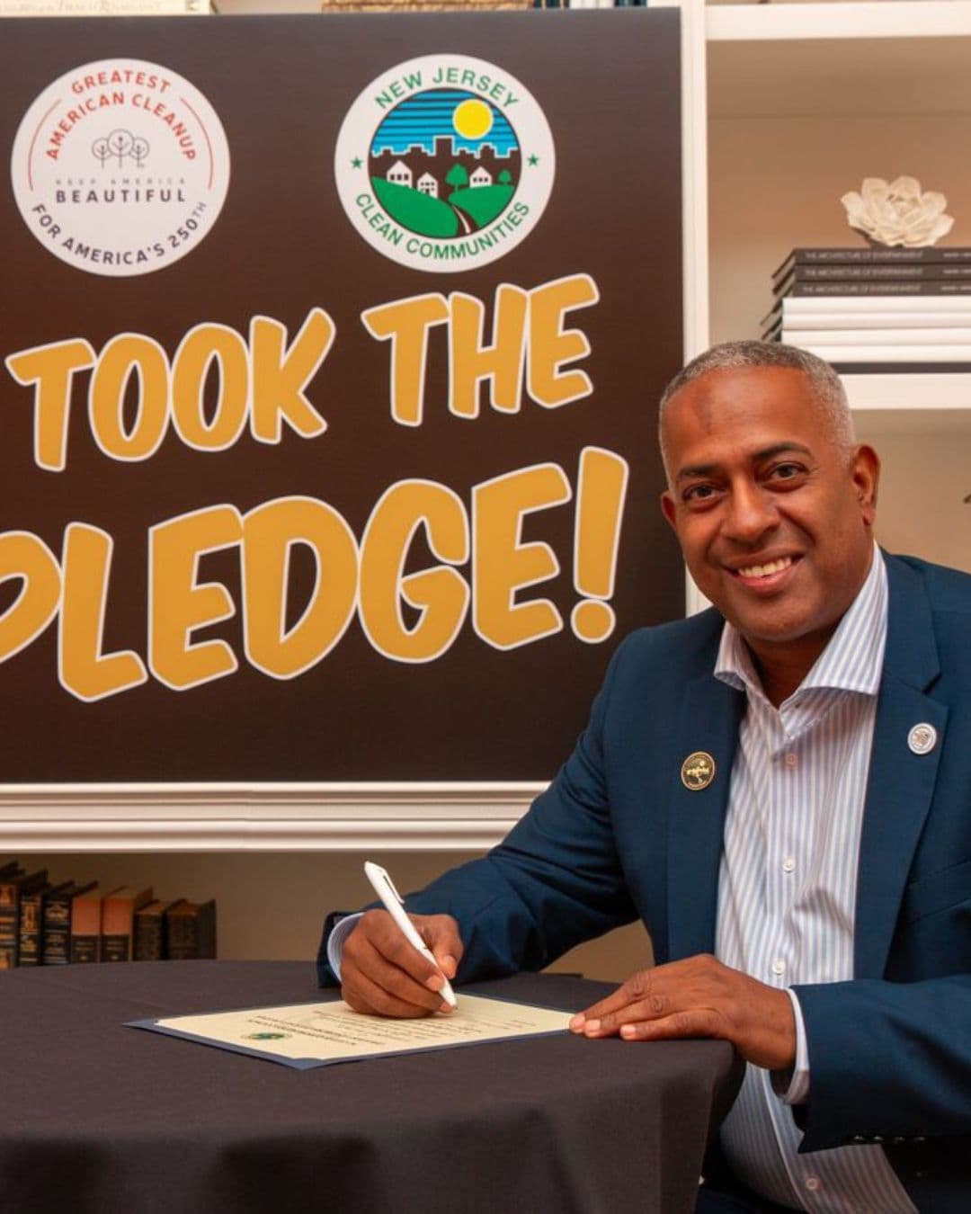 A man in a navy blazer and striped shirt smiles at the camera while signing a certificate on a dark cloth-covered table. Behind him a brown board reads TOOK THE PLEDGE! beside a Greatest American Cleanup for America's 250th roundel and a New Jersey Clean Communities seal.