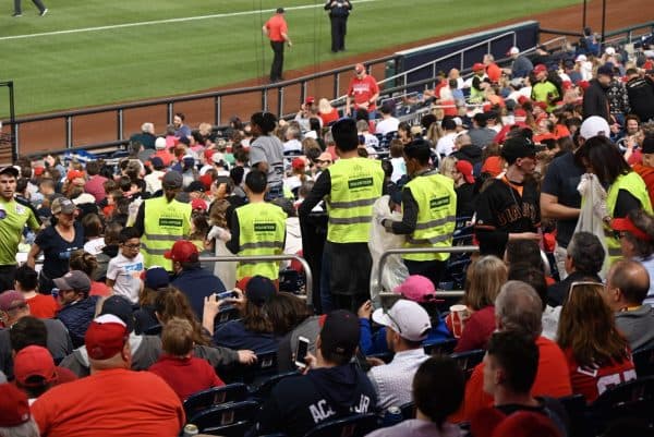 Several people in yellow VOLUNTEER vests move through packed baseball stadium seating collecting waste in bags, surrounded by spectators in red caps and shirts with the green outfield visible beyond.