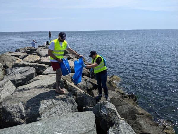 Two men work across a granite breakwater beside calm blue-grey sea, one in a yellow hi-vis vest holding open a blue rubbish bag and a grabber, the other crouching to reach litter between the boulders. A third figure fishes further out on the rocks.