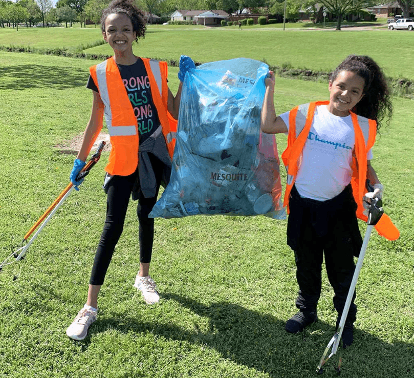 Two smiling girls in orange safety vests stand on a green park lawn holding up a large clear blue rubbish sack printed MESQUITE and packed with cans and bottles, each with a long litter grabber in her other hand.