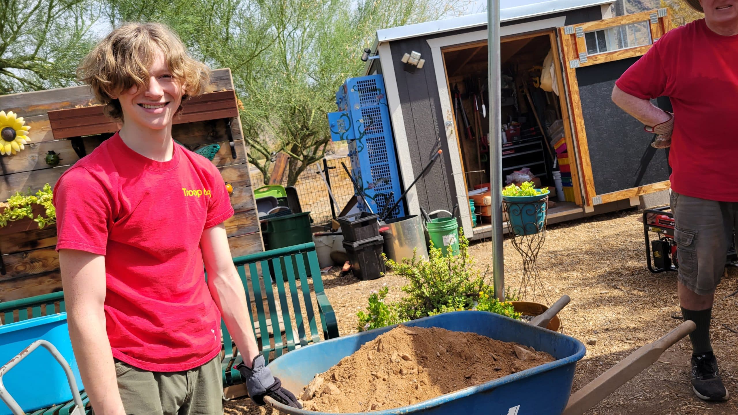 Smiling teenager in a red troop T-shirt and work gloves stands beside a blue wheelbarrow heaped with dirt in a desert community garden