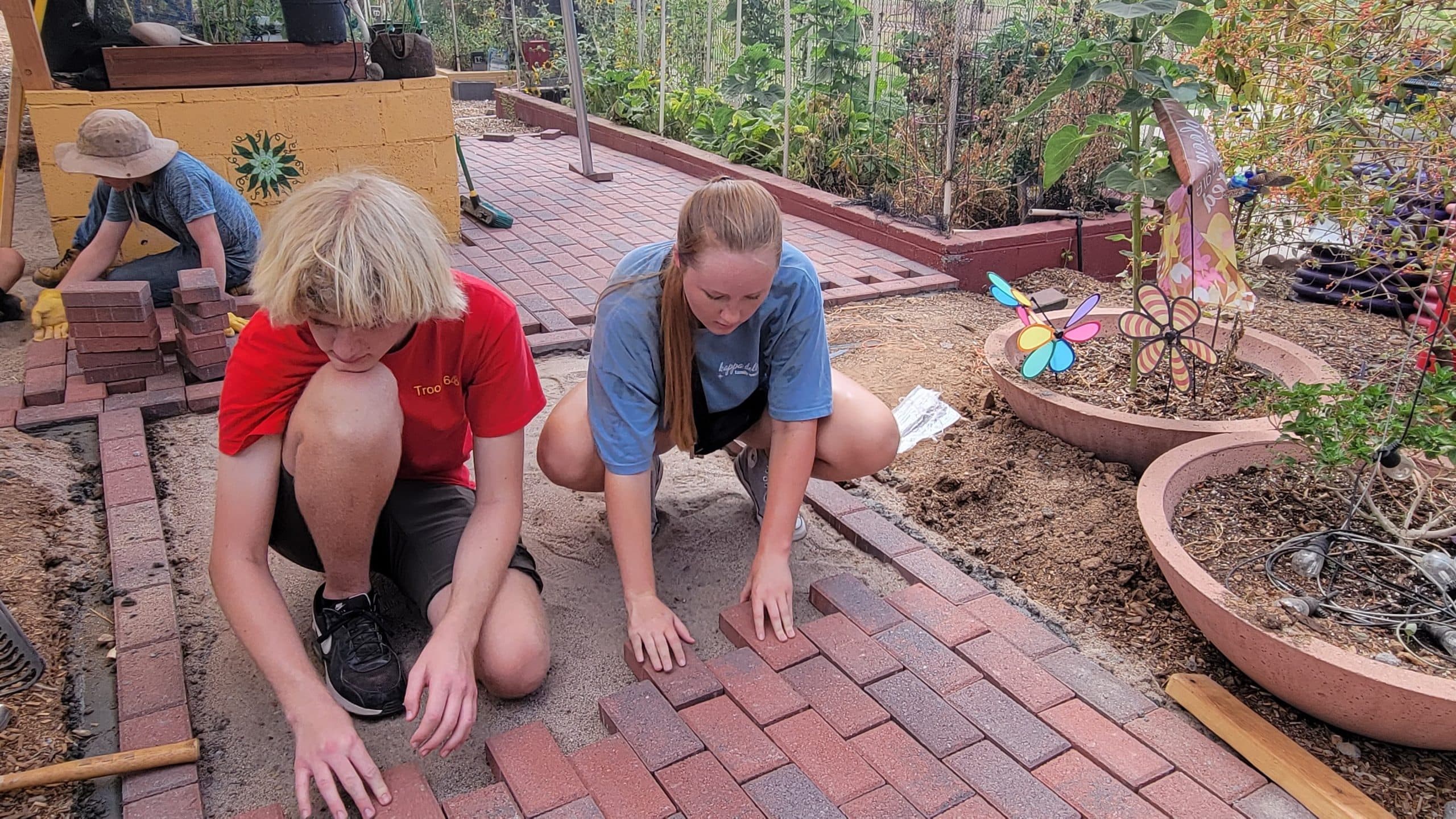 Two teenagers kneel on sand setting red clay pavers into a new garden path, pinwheel flowers and concrete planters nearby