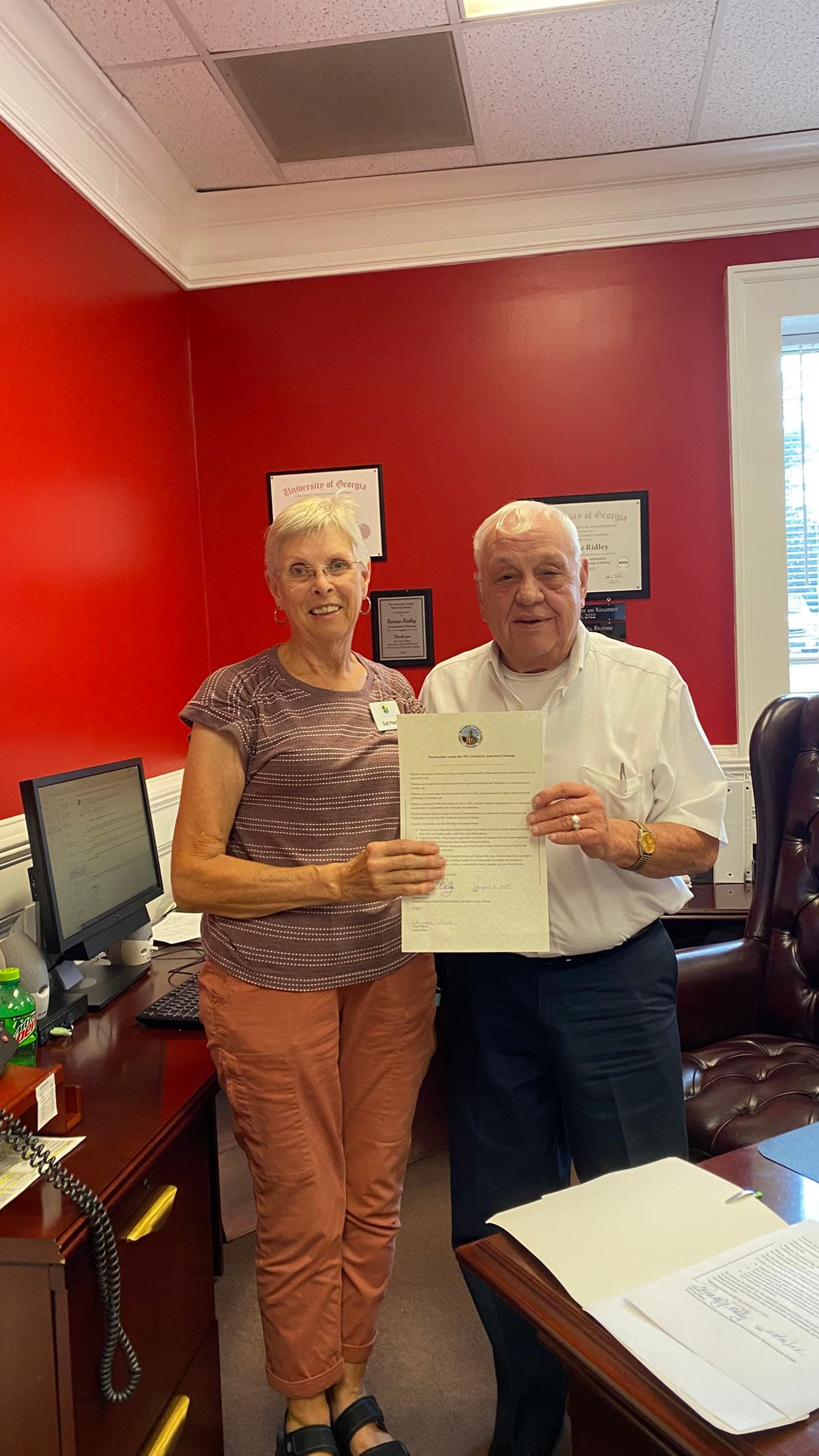 An older woman in a striped mauve top and an older man in a white short-sleeved shirt stand together in a red-walled office holding a printed Declaration Action for The Great(est) American Cleanup between them, framed University of Georgia certificates on the wall behind.