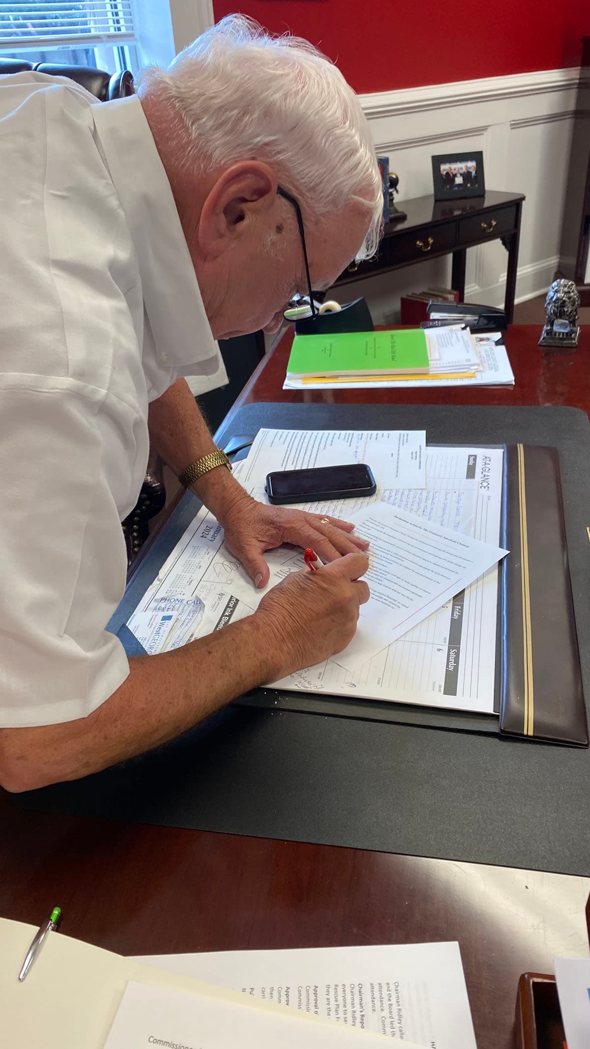 A white-haired man in a short-sleeved white shirt and reading glasses leans over a desk blotter, signing a printed document with a red pen. A mobile phone, desk calendar and stacks of paperwork cover the desk around him.