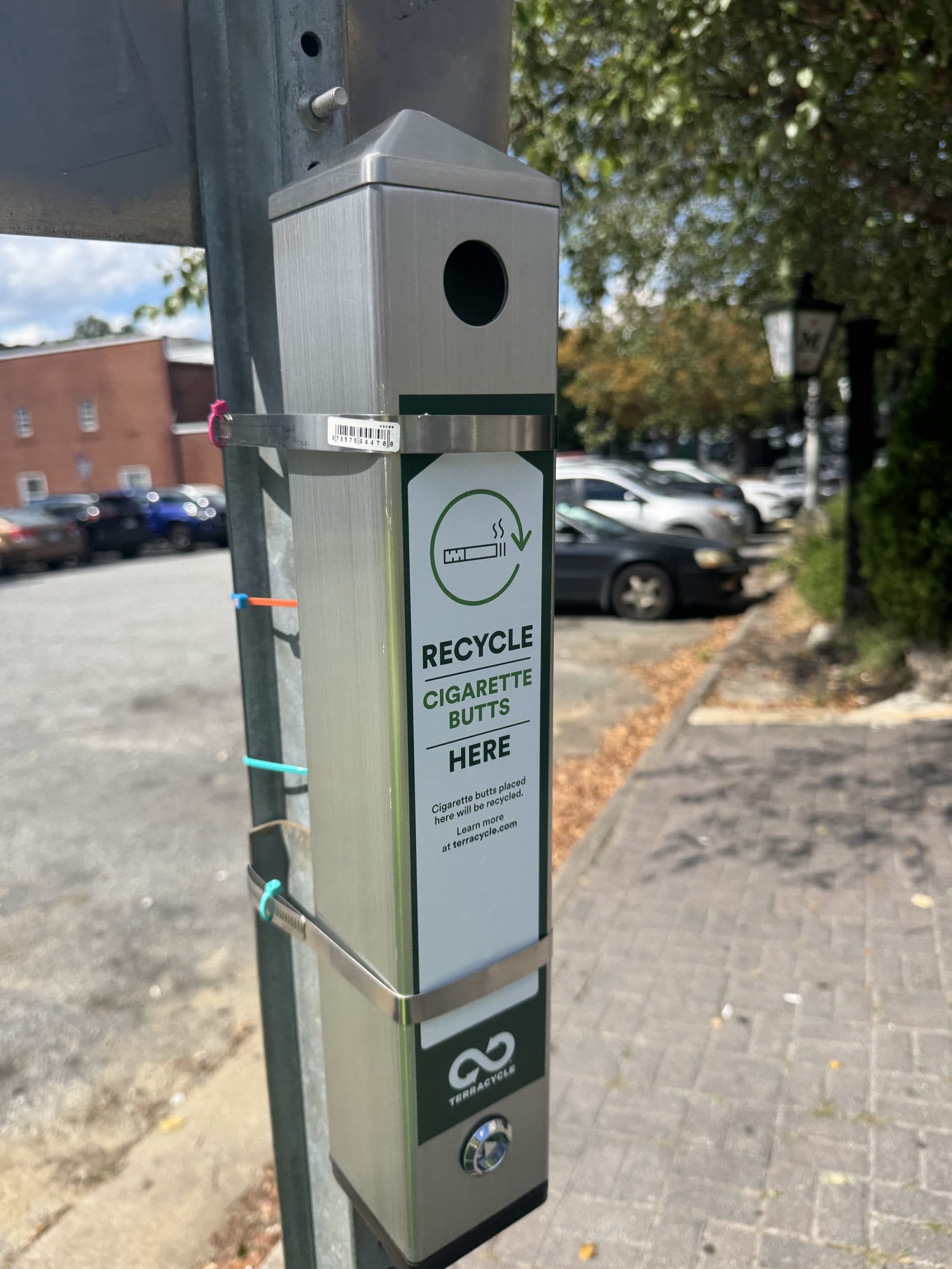 A brushed steel cigarette-butt receptacle strapped with metal bands to a grey pole on a brick pavement. Its label reads RECYCLE CIGARETTE BUTTS HERE, with a cigarette-in-a-circle icon, the note that butts placed here will be recycled, and a TerraCycle logo. A car park and street lamp sit out of focus behind.