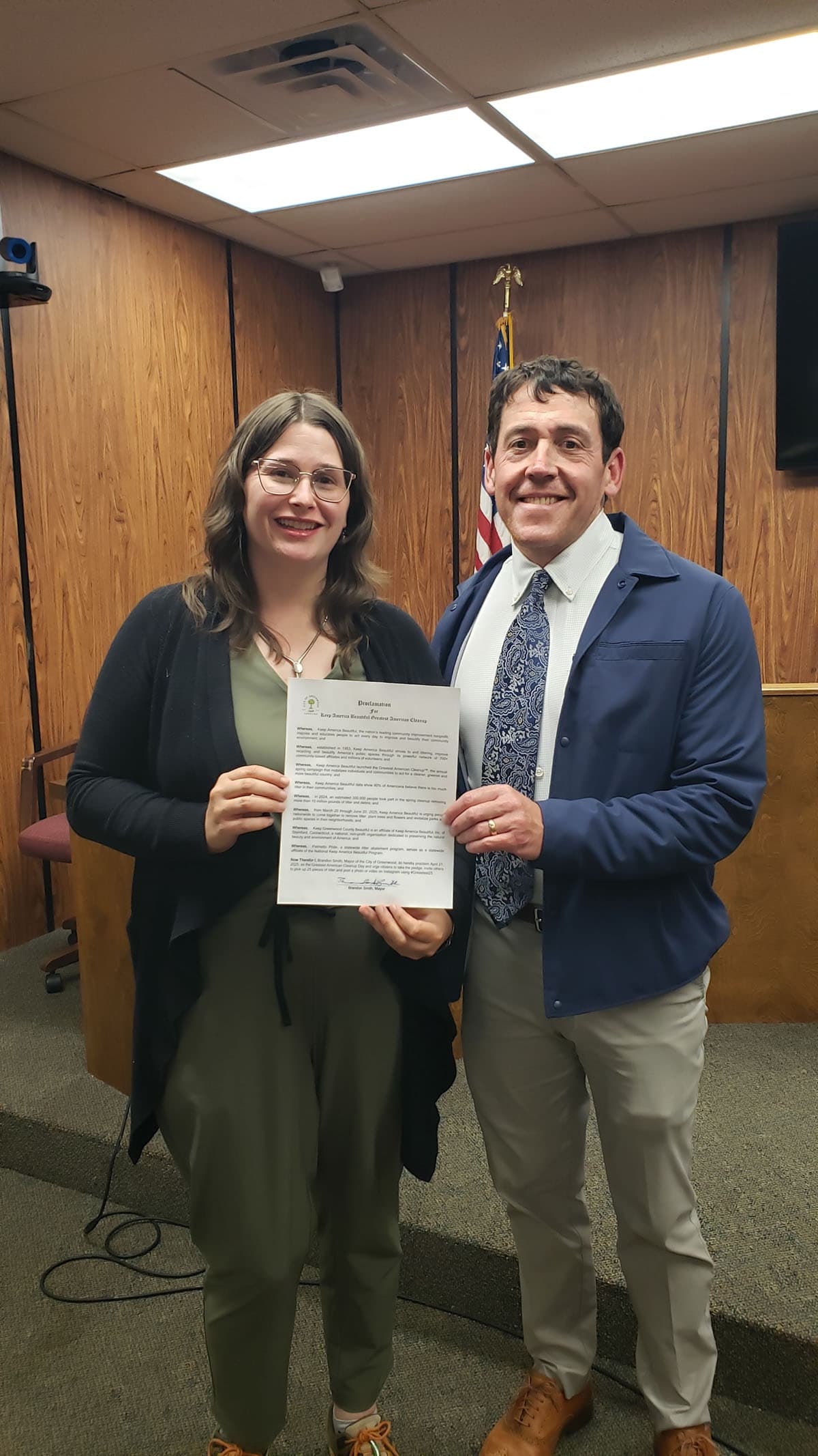 A woman in a dark cardigan and glasses and a man in a navy jacket and paisley tie stand in a wood-panelled council chamber, holding a printed Proclamation between them. A US flag and a lectern stand behind.