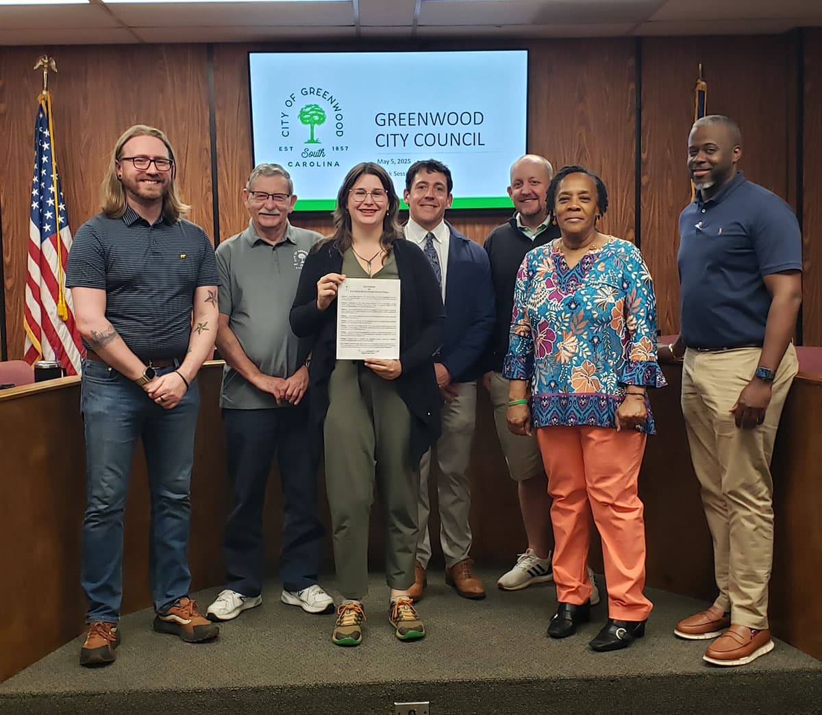 Seven people line up on a council chamber dais, the woman at centre holding a printed proclamation. The screen behind them shows a green tree emblem and the words CITY OF GREENWOOD, SOUTH CAROLINA and GREENWOOD CITY COUNCIL, with a US flag to the left.