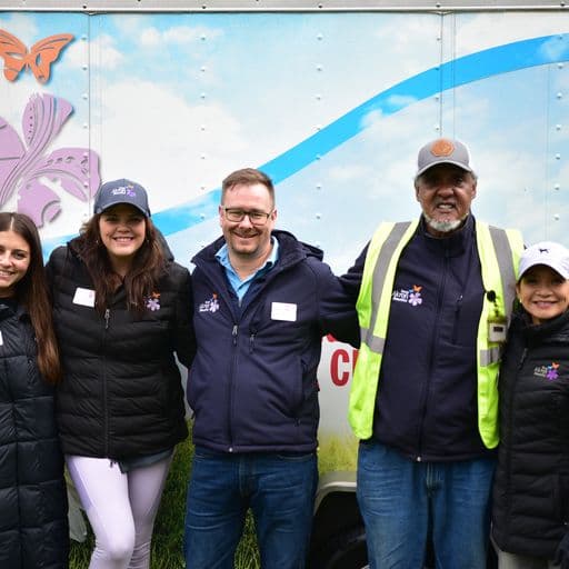 Five volunteers in dark jackets and caps stand arm in arm in front of a box truck painted with a butterfly, sky and grass mural