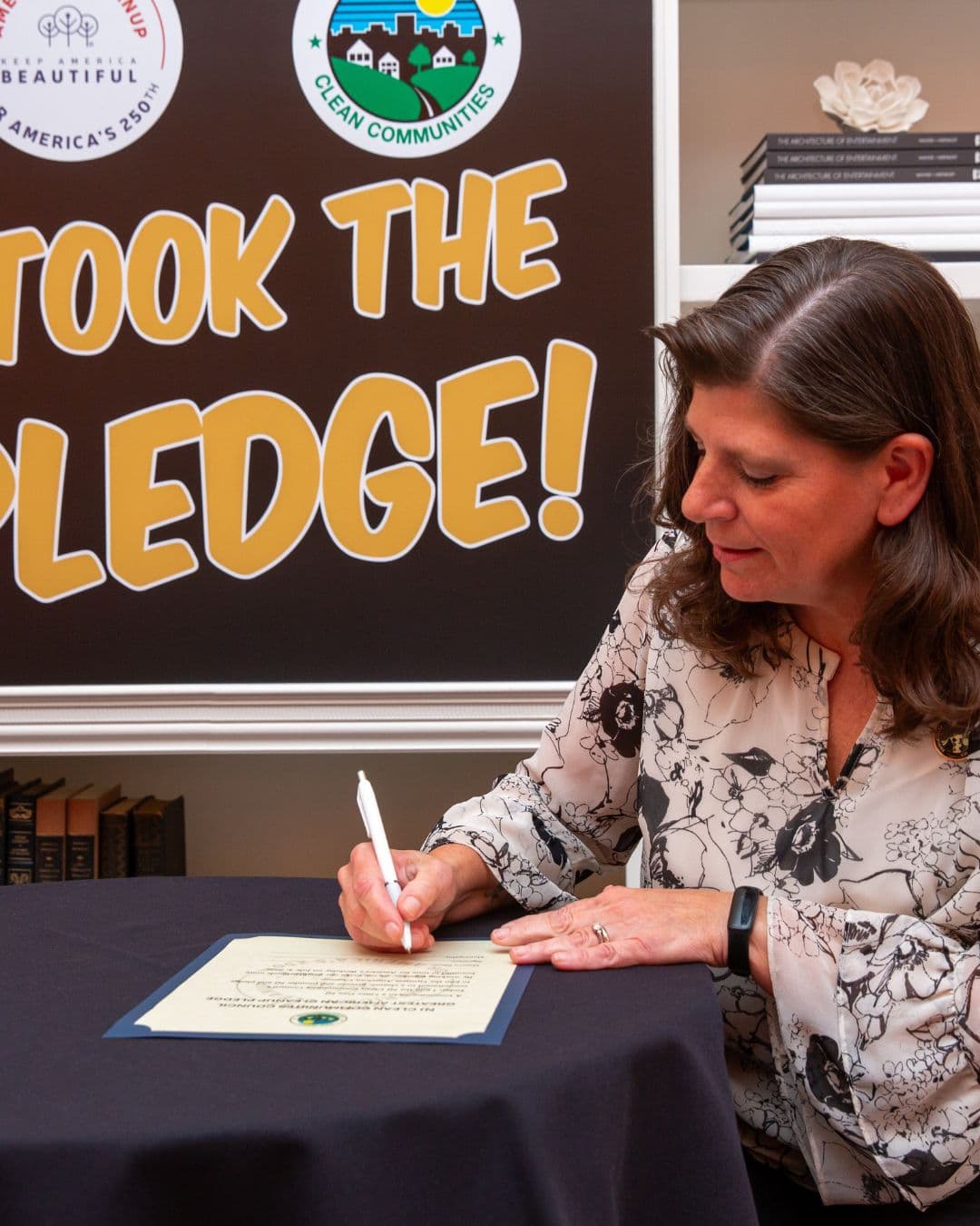 A woman with shoulder-length brown hair in a black-and-white floral blouse signs a certificate laid on a navy cloth table. Behind her a large brown board reads TOOK THE PLEDGE! next to a Clean Communities roundel and bookshelves.