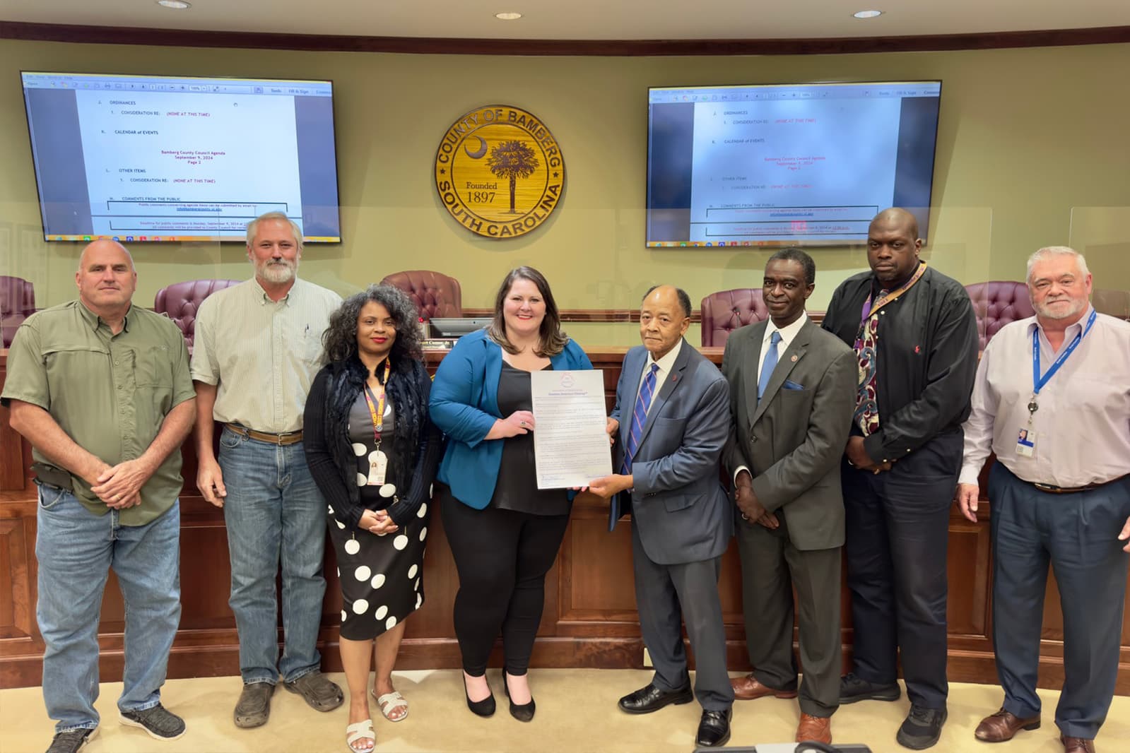 Nine people stand in a wood-panelled council chamber beneath a gold seal reading COUNTY OF BAMBERG, SOUTH CAROLINA, FOUNDED 1897. A woman in a teal blazer and a man in a grey suit hold a printed proclamation between them, with agenda pages displayed on screens either side.