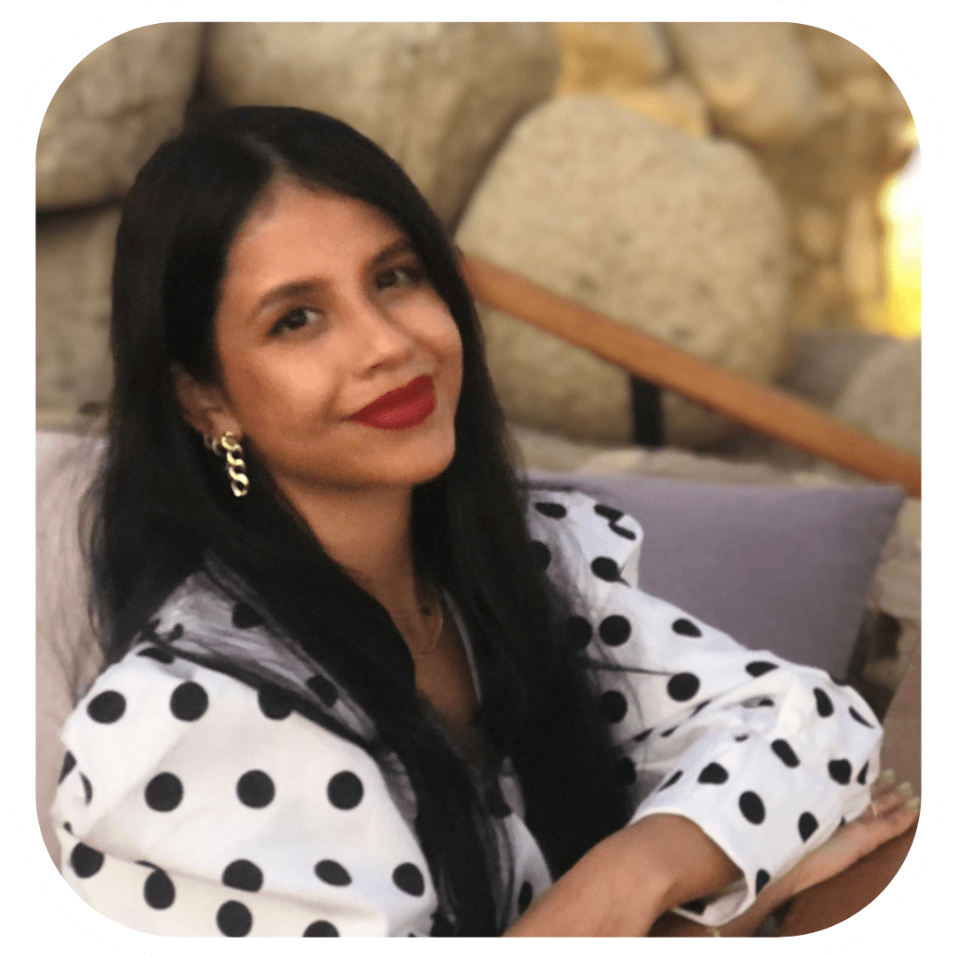 Portrait in a rounded white frame of a woman with long straight black hair and red lipstick, wearing a white jacket with large black polka dots and a gold chain earring, reclining on a cushioned seat in front of a pale stone wall.