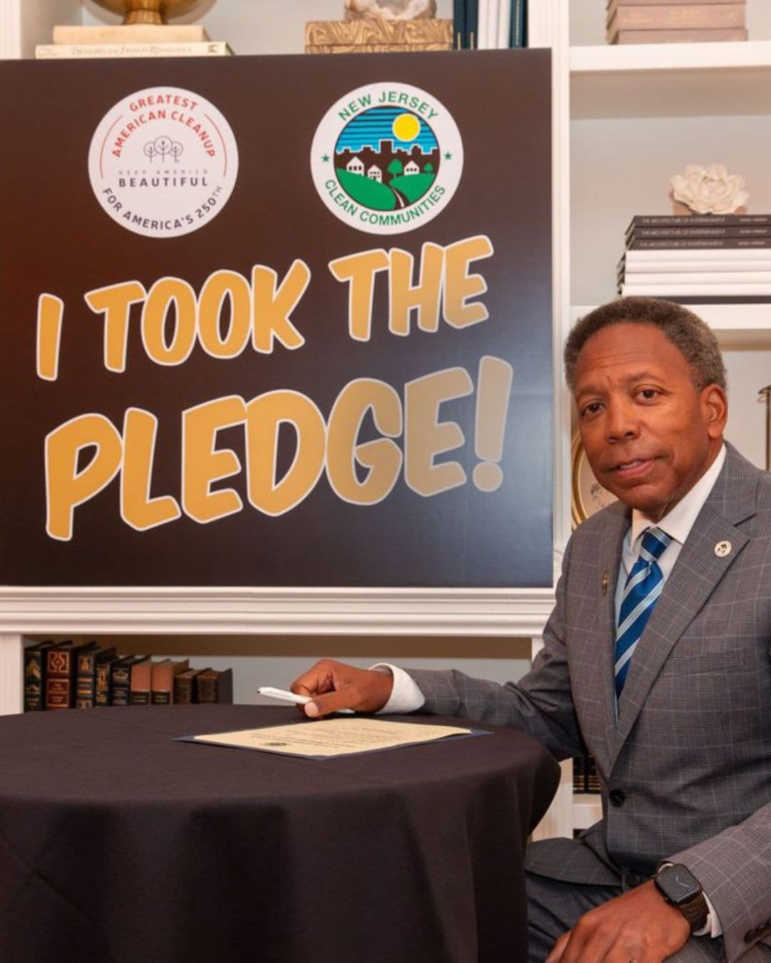 A man in a grey checked suit and blue striped tie sits at a round table draped in dark cloth with a signed certificate in front of him. A large board behind reads I TOOK THE PLEDGE! beside a Greatest American Cleanup for America's 250th badge, set against white bookshelves.