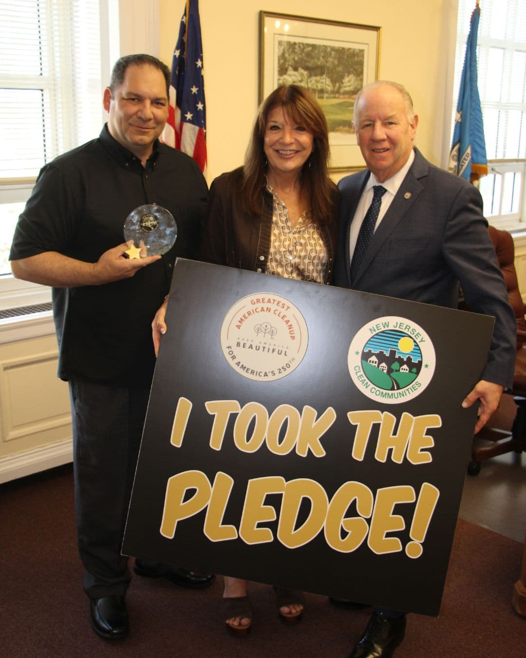 Three people stand in an office with US and state flags behind them, holding a large black board that reads I TOOK THE PLEDGE! with a Greatest American Cleanup badge and a New Jersey Clean Communities seal. The man on the left also holds a round glass award with a gold star.