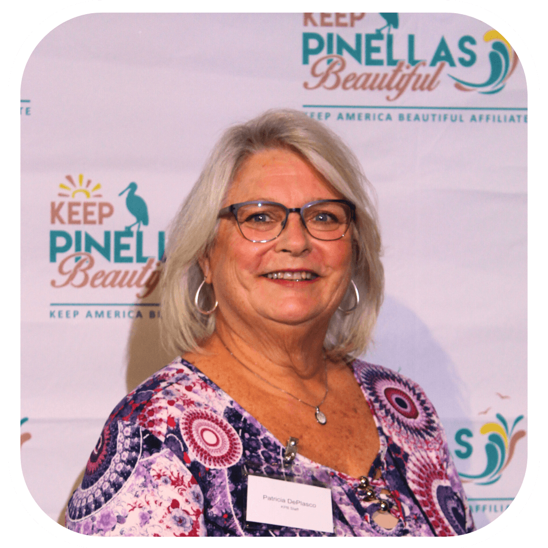 Portrait of a smiling woman with silver-blonde shoulder-length hair, hoop earrings and dark-rimmed glasses in a purple and magenta patterned top. Her name badge reads Patricia DePlasco, and a Keep Pinellas Beautiful backdrop with a pelican logo fills the wall behind.