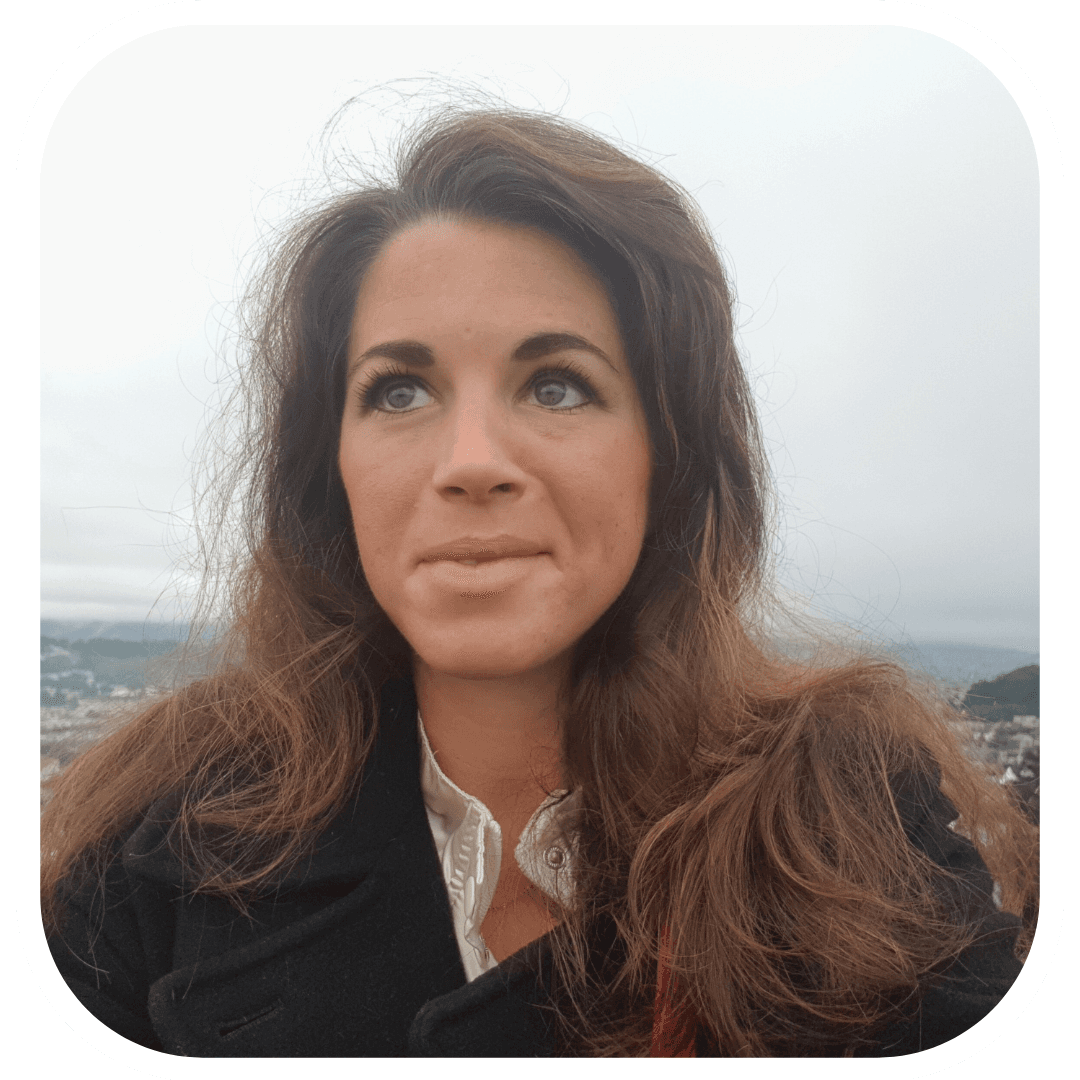 Portrait of a woman with long wavy chestnut hair in a black wool coat over a cream ruffled blouse, looking up and away from the camera. A hazy view of distant hills and rooftops sits behind her under a flat white sky.