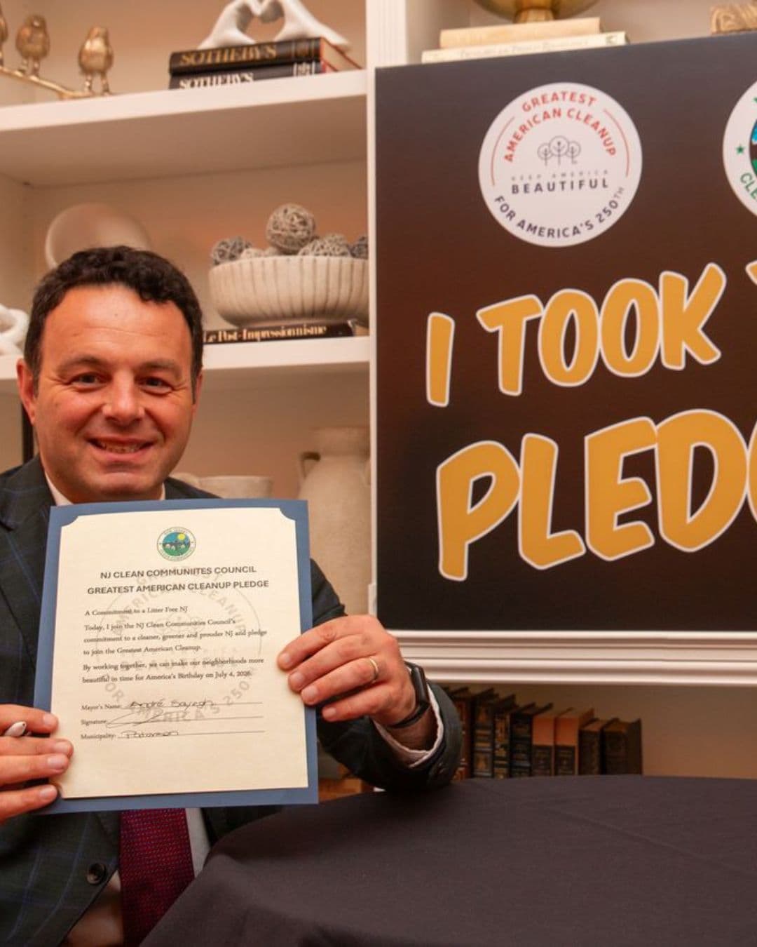 A man in a blue plaid suit holds up a completed certificate headed NJ CLEAN COMMUNITIES COUNCIL, GREATEST AMERICAN CLEANUP PLEDGE, a commitment to a litter-free NJ, with the mayor and municipality lines filled in by hand. A large I TOOK THE PLEDGE board stands behind him beside shelves of books and ornaments.