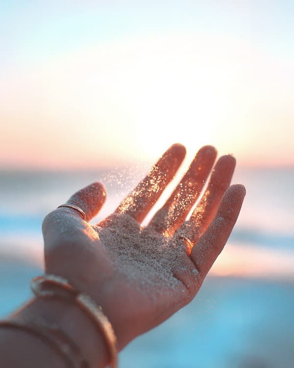 An open palm held up against a bright low sun at the coast, pale sand pouring between the fingers and glittering in the light. Stacked rings and bangles are visible on the hand and wrist; sea and sky blur behind.