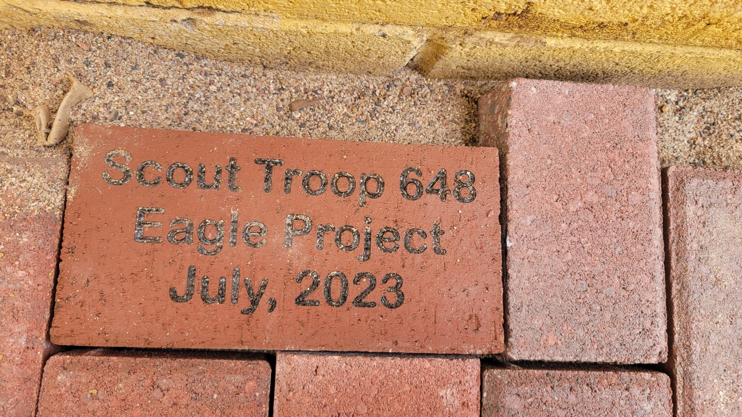 Engraved red paver reading Scout Troop 648 Eagle Project July 2023 set into a new brick path against a yellow block wall