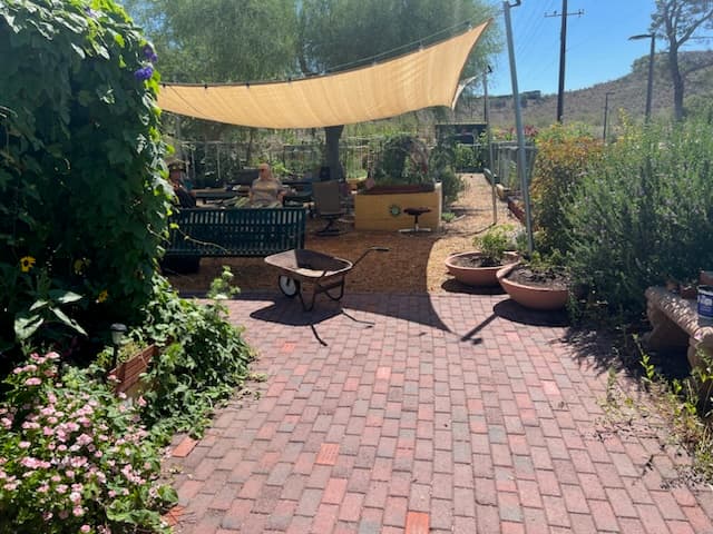 Finished brick patio in a desert community garden with an empty wheelbarrow, tan shade sail, potted plants and pink flowers, hills behind