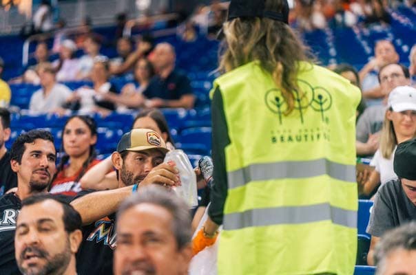 Seen from behind, a person in a lime hi-vis vest printed with the Keep America Beautiful three-trees mark and the word BEAUTIFUL holds a clear sack open in a stadium aisle while a seated fan in a camouflage cap drops in an empty plastic bottle. Rows of spectators fill the frame.