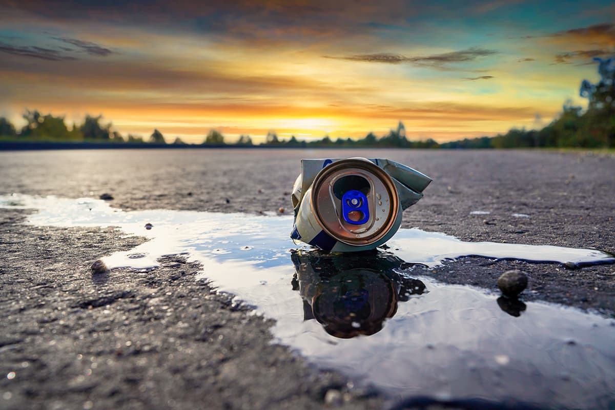 A crushed silver and blue drinks can lying on its side in a shallow puddle on coarse asphalt, mirrored in the water. The horizon behind glows orange and gold at sunset above a distant treeline.