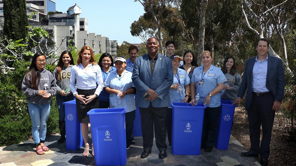 Twelve people pose on a tiled campus terrace with four tall blue recycling bins carrying the Keep America Beautiful tree logo. Several wear pale blue uniform shirts, two wear UC San Diego sweatshirts, and a man in a grey suit stands at the centre. Eucalyptus trees and a concrete building rise behind.