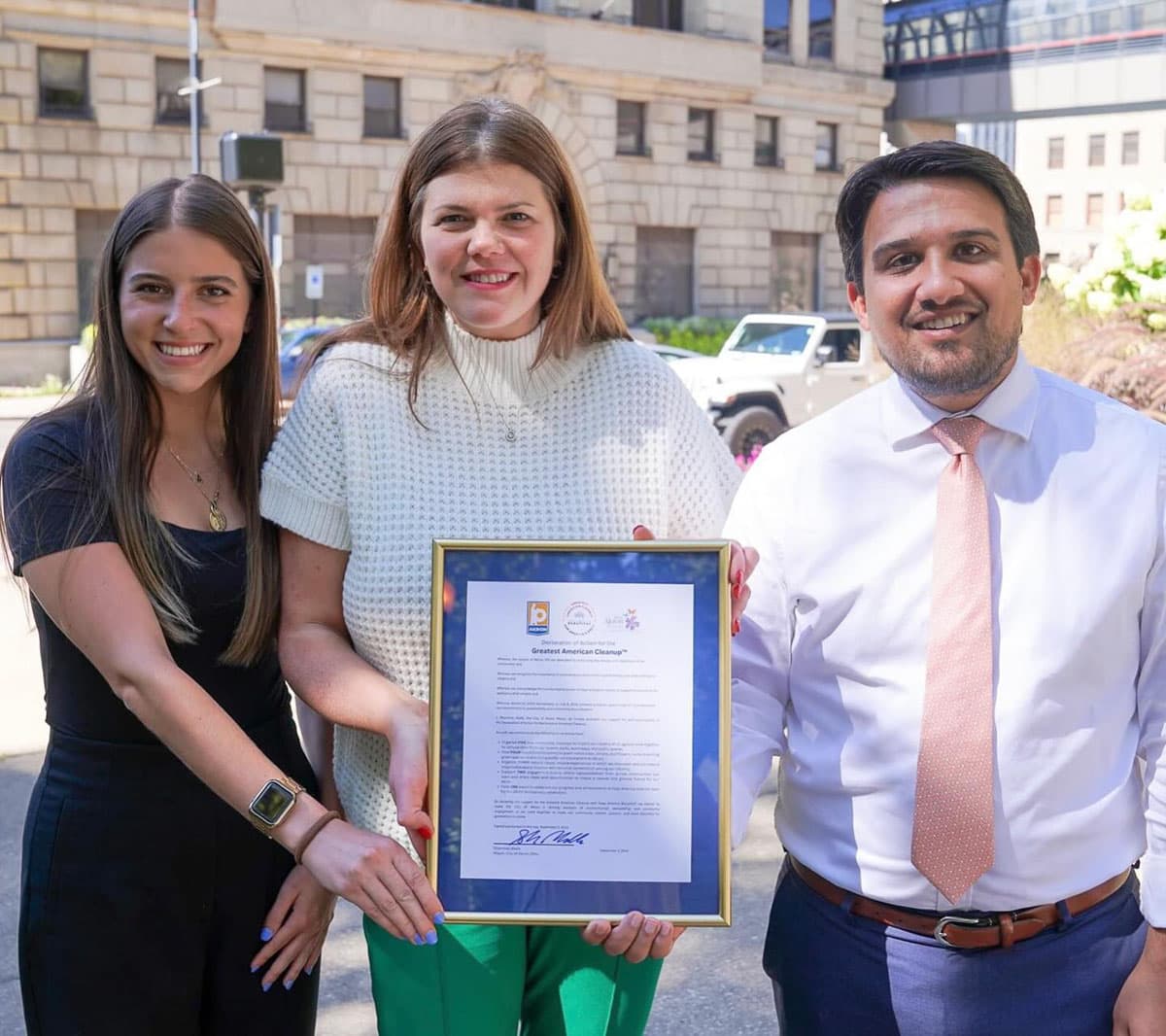 Three people stand shoulder to shoulder outside a stone civic building holding a gold-framed printed Declaration of Action for the Greatest American Cleanup. A woman in a cream knitted top holds the frame, flanked by a woman in black and a man in a white shirt and pink tie.