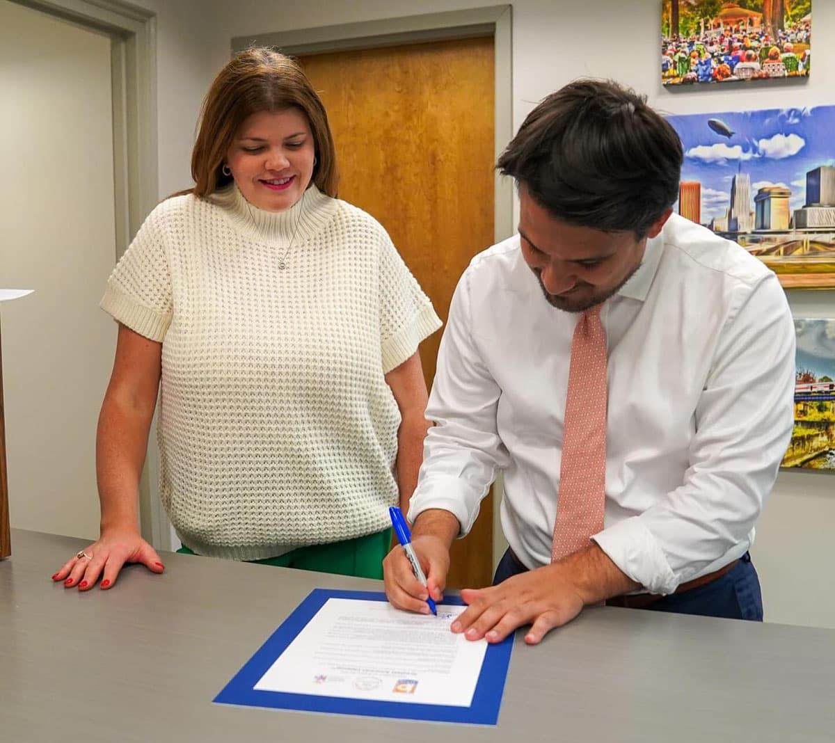 A man in a white shirt and pink tie leans over a grey counter to sign a proclamation mounted on blue card with a blue pen, while a woman in a cream knitted top stands beside him watching. Framed cityscape paintings hang on the wall behind.