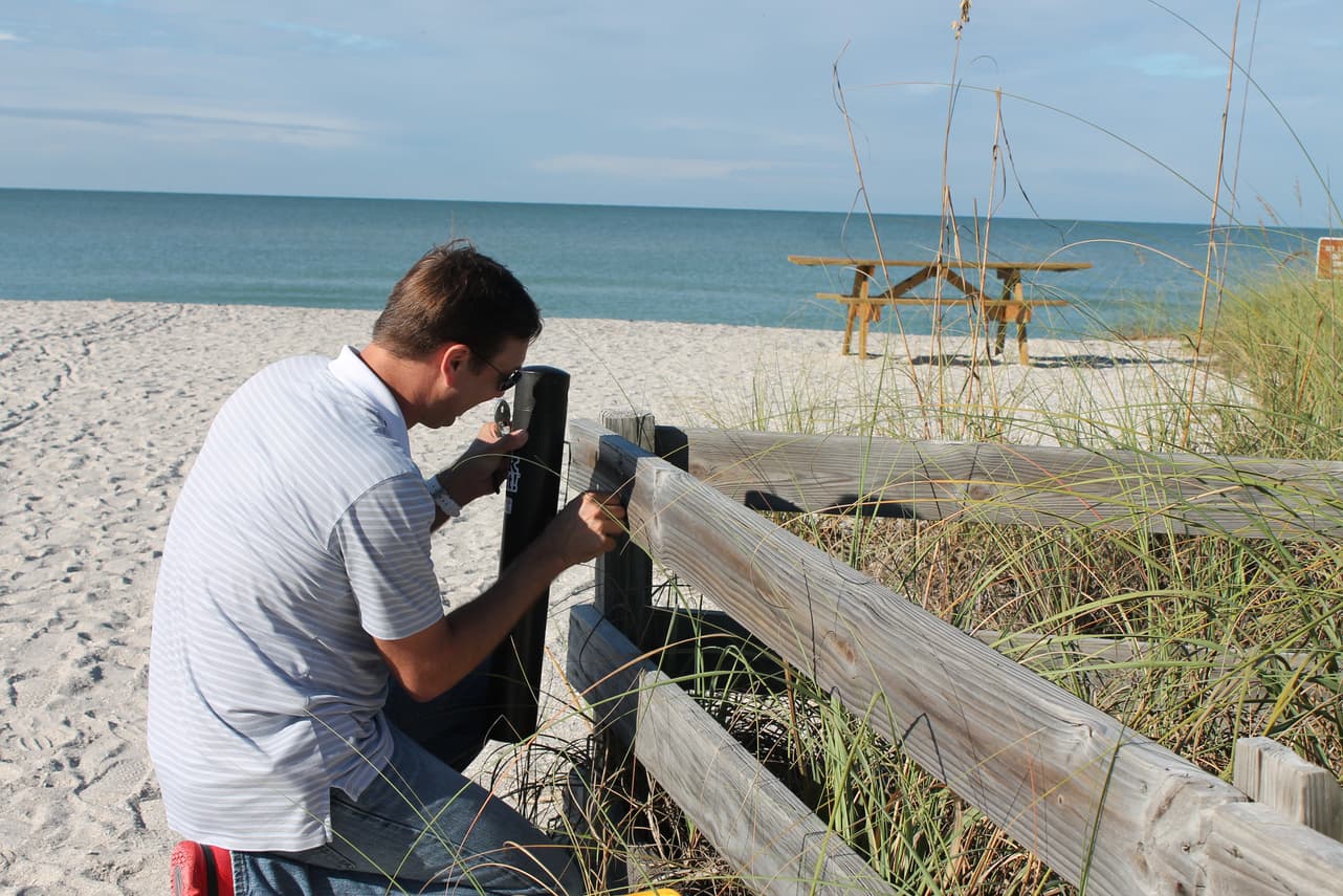 A man in a white striped polo kneels beside a weathered split-rail fence at the top of a beach, fitting a black cylindrical receptacle to a post. Sea oats and dune grass fill the right of the frame, with white sand, a wooden picnic table and blue-green ocean behind.