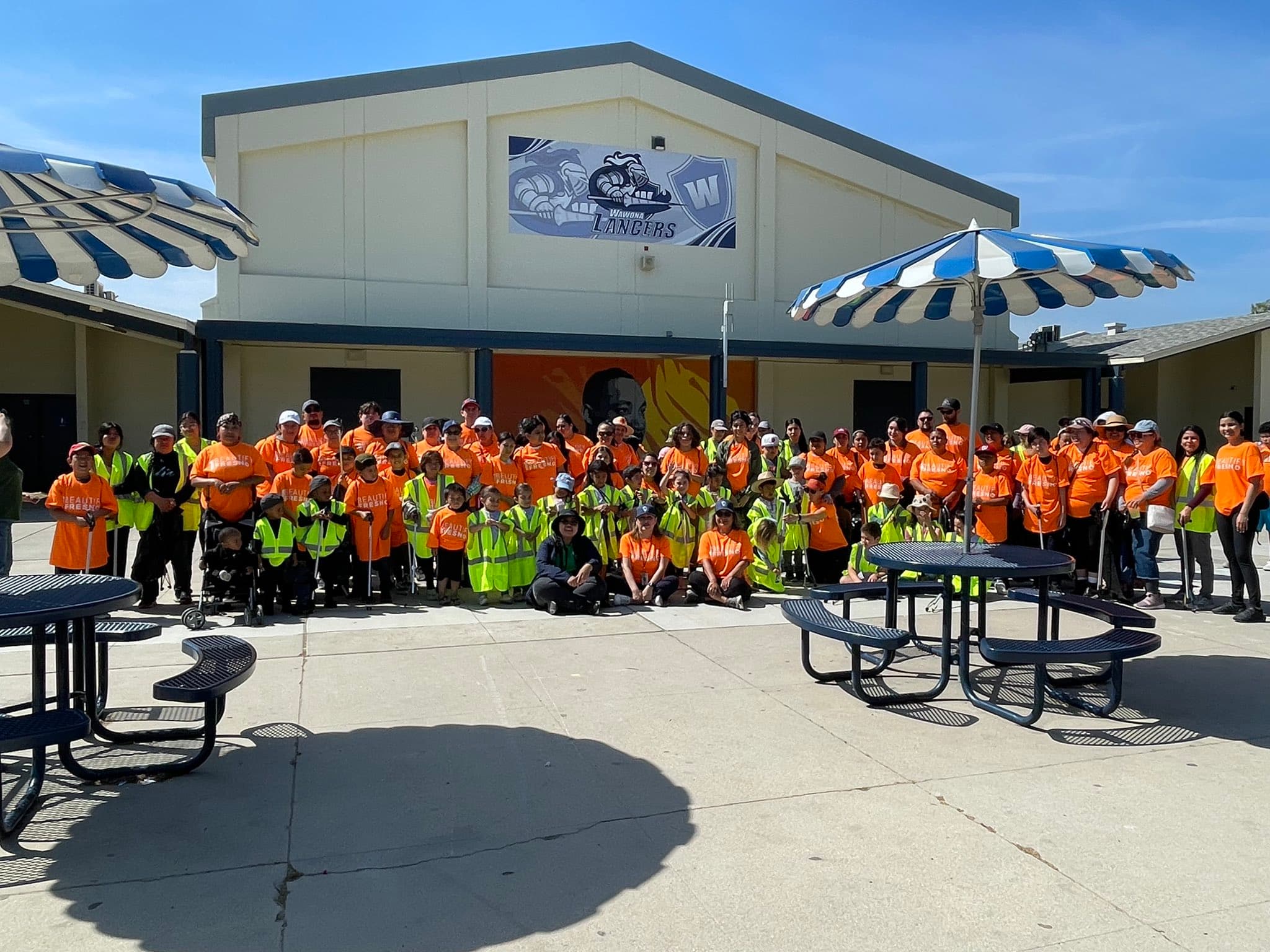 About a hundred volunteers in orange BEAUTIFY FRESNO shirts and high-visibility vests pose with litter grabbers outside a school gym