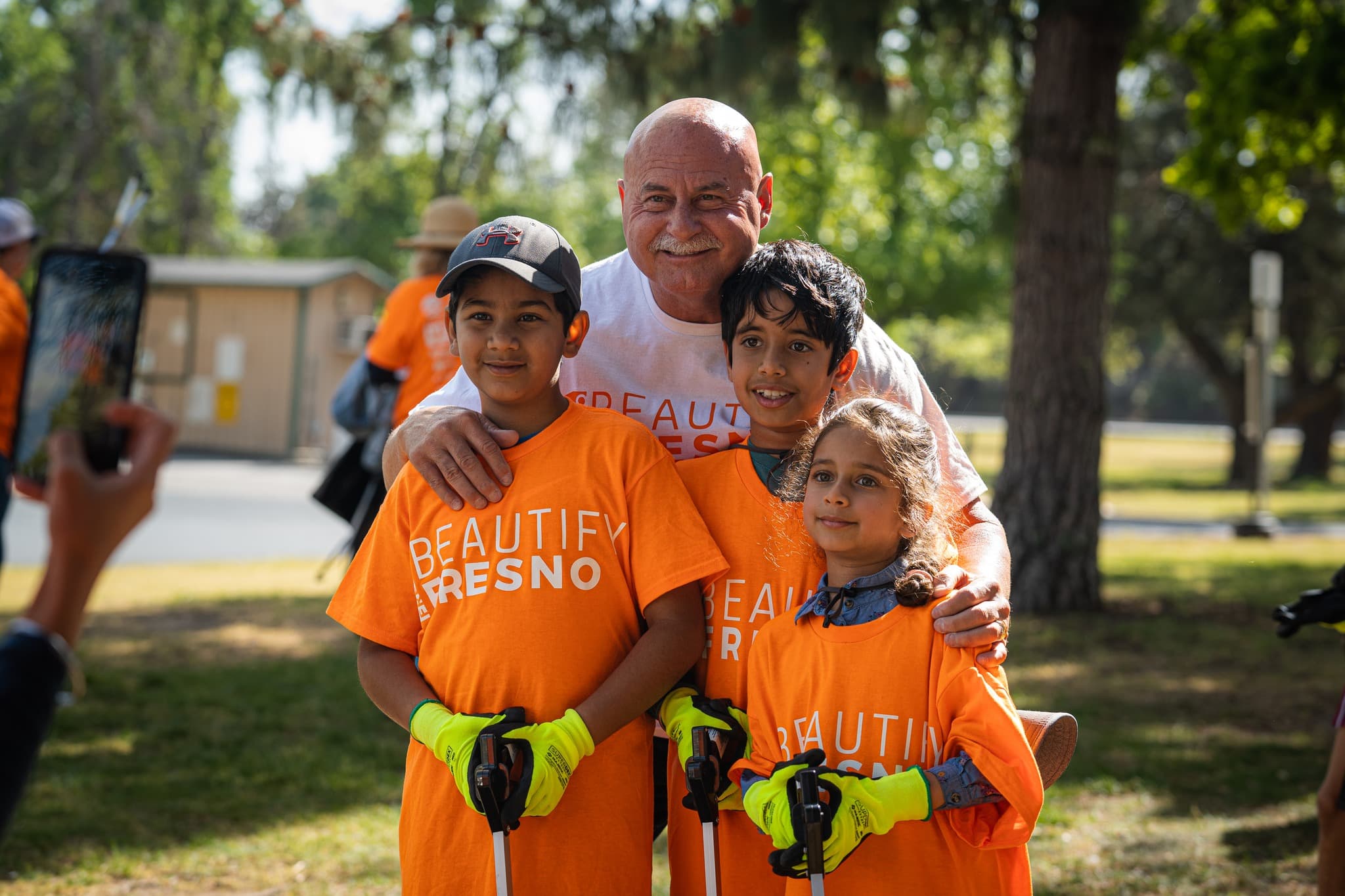 Bald man in a white Beautify Fresno shirt with his arms around three children in orange shirts and lime gloves holding litter grabbers