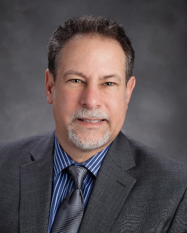 Studio headshot of a man with short greying hair and a white goatee, wearing a grey checked jacket, blue pinstripe shirt and grey patterned tie, against a mottled grey backdrop.