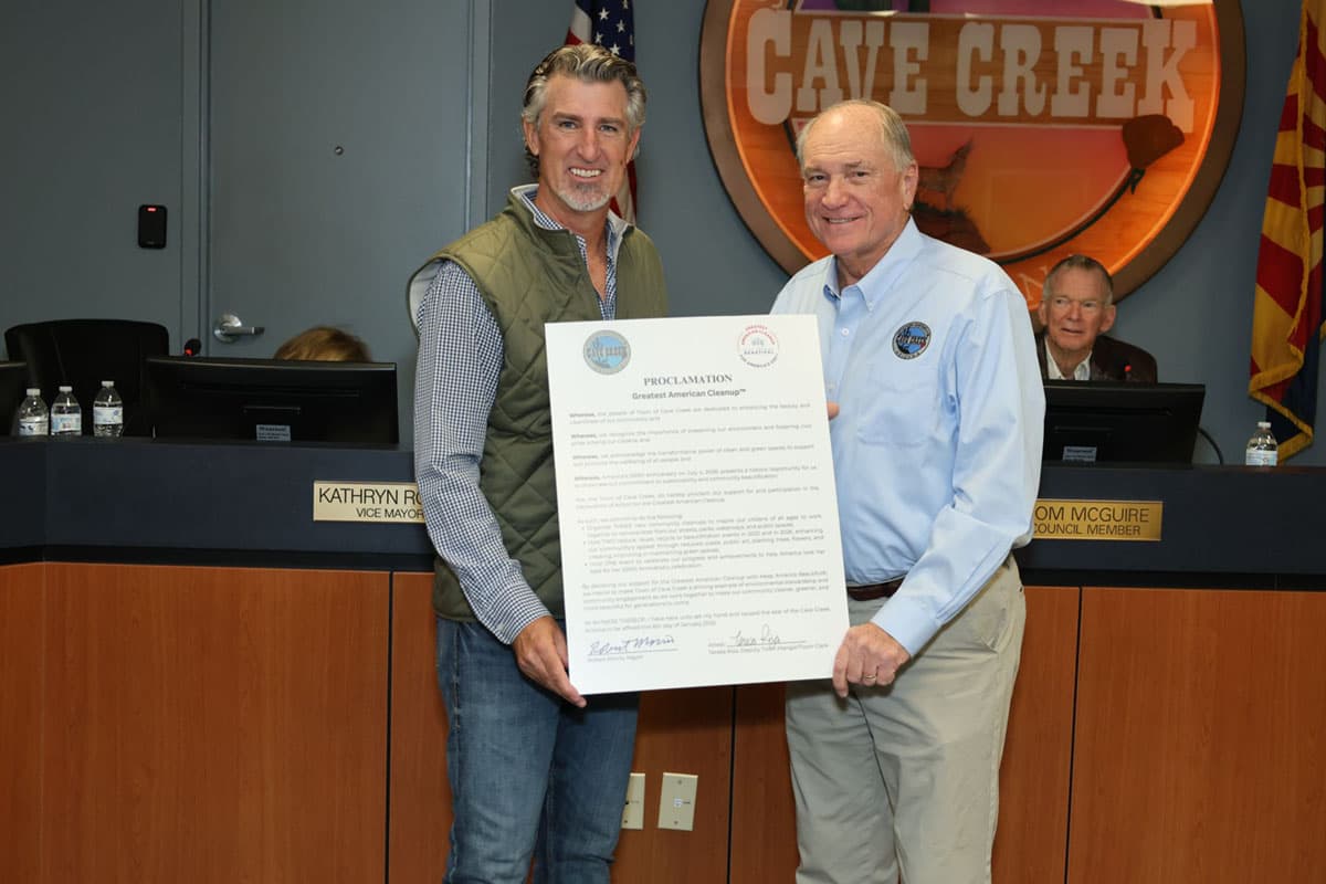 Two men stand in a council chamber holding a large printed PROCLAMATION for the Greatest American Cleanup between them: one in a green gilet over a checked shirt and jeans, the other in a pale blue shirt with a round town badge. A carved CAVE CREEK sign hangs behind, and desk nameplates read VICE MAYOR and TOM MCGUIRE, COUNCIL MEMBER.