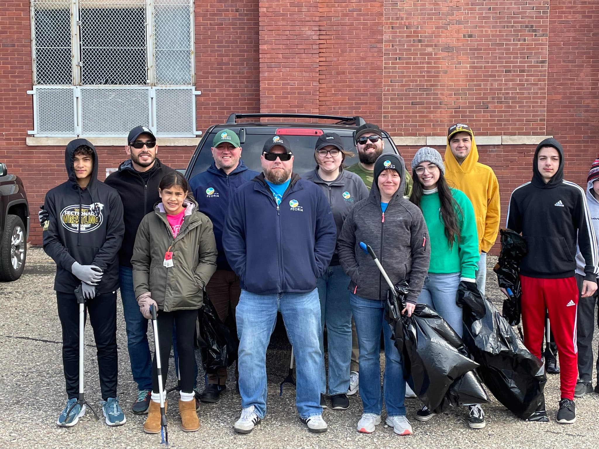 Bundled-up volunteers with litter grabbers and black bags stand in front of a brick building on an overcast day