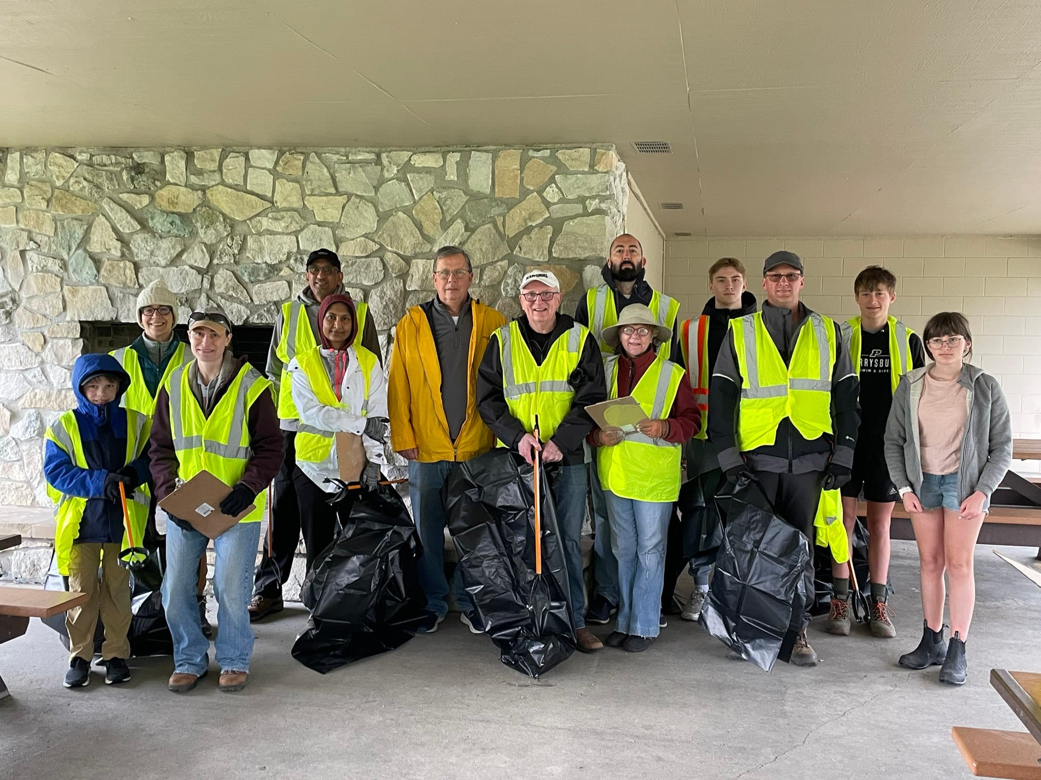 Fifteen volunteers in lime safety vests hold black trash bags and clipboards inside a stone-fireplace park shelter