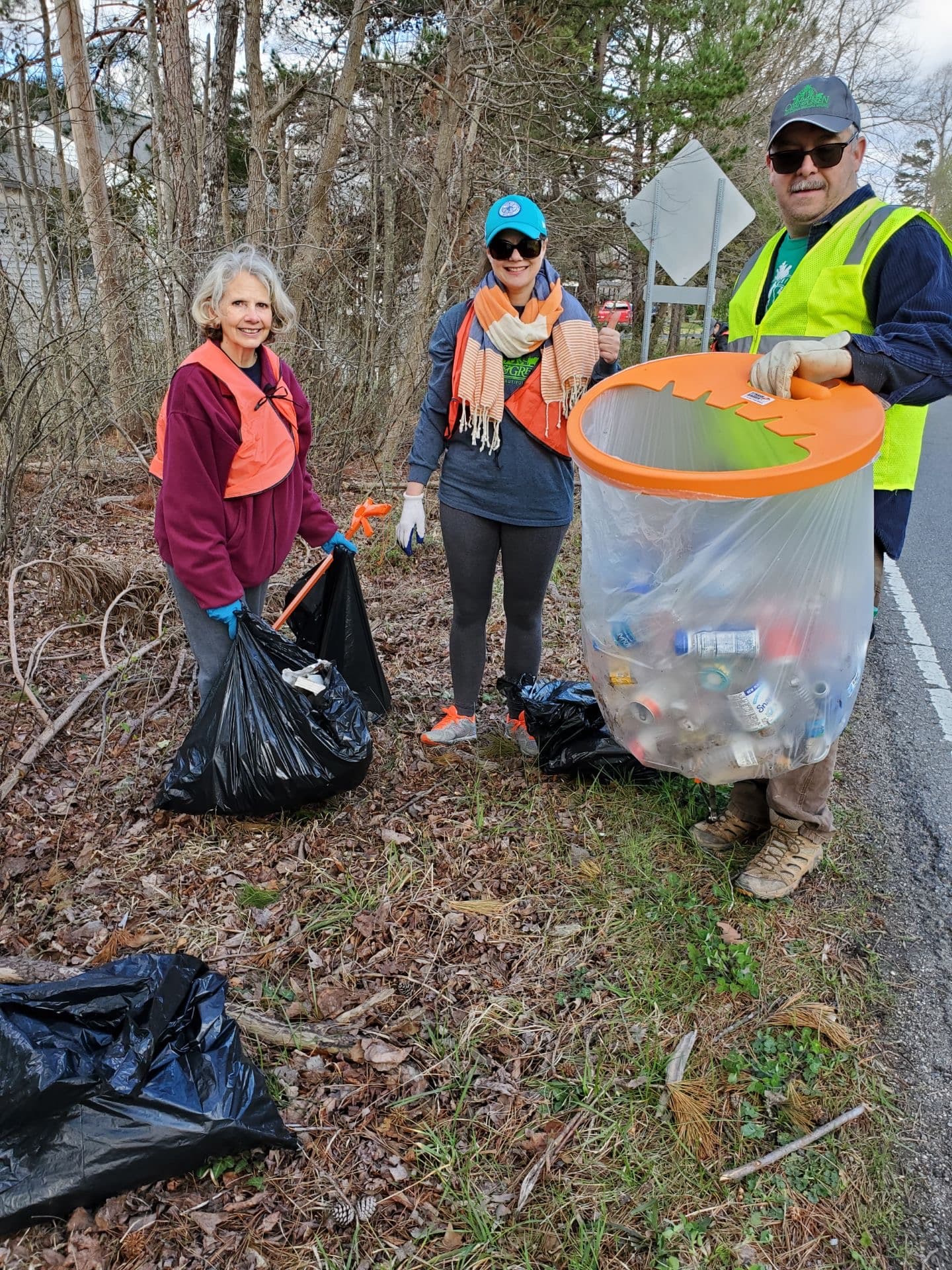 Two women with black bags and a man in a lime vest holding a clear bag full of plastic bottles on a wooded roadside verge