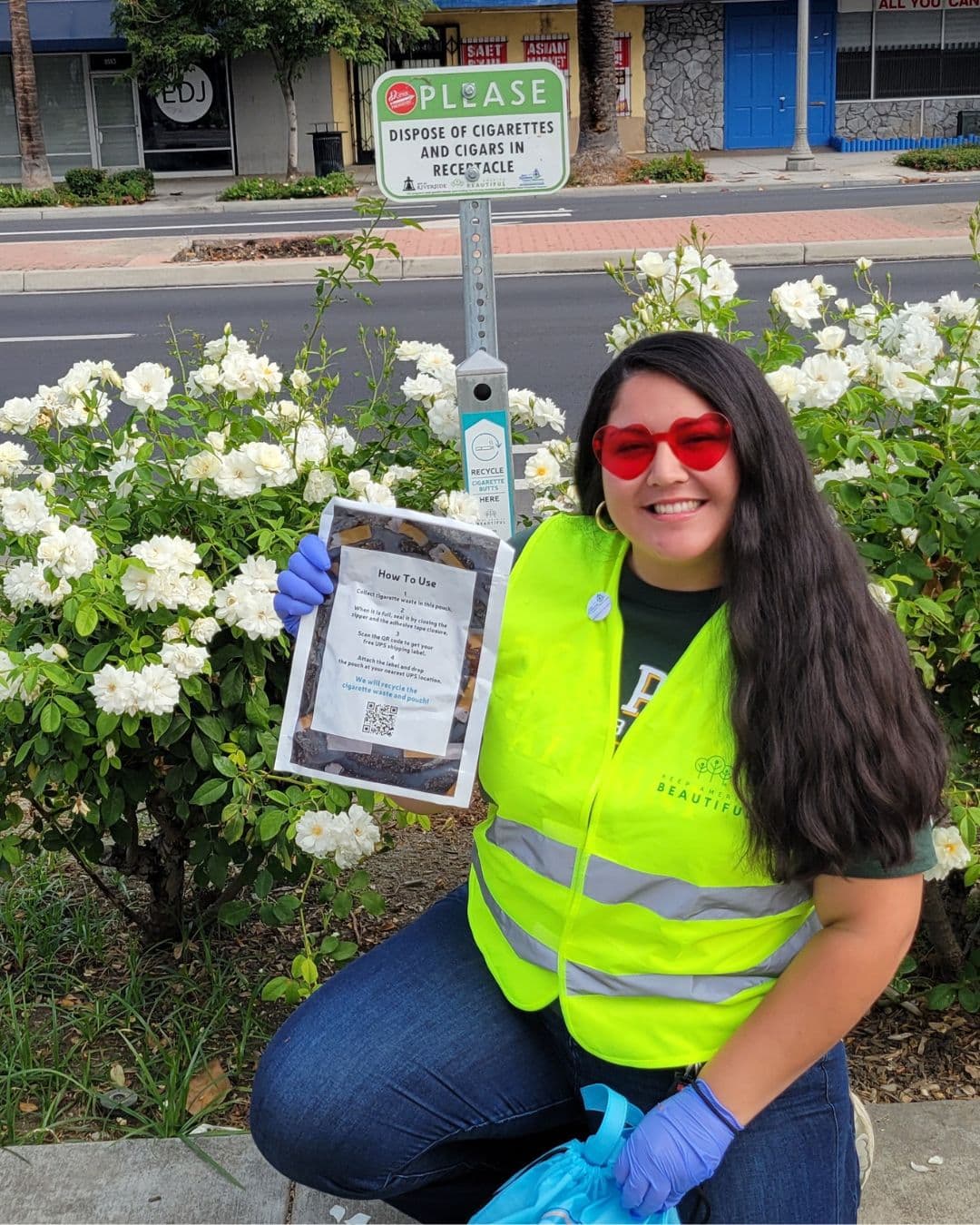 A smiling woman in a yellow hi-vis vest and red heart-shaped sunglasses kneels by a white rose bush on a city street, holding up a clear pouch of cigarette butts labelled How To Use. A pole sign behind her reads PLEASE DISPOSE OF CIGARETTES AND CIGARS IN RECEPTACLE.